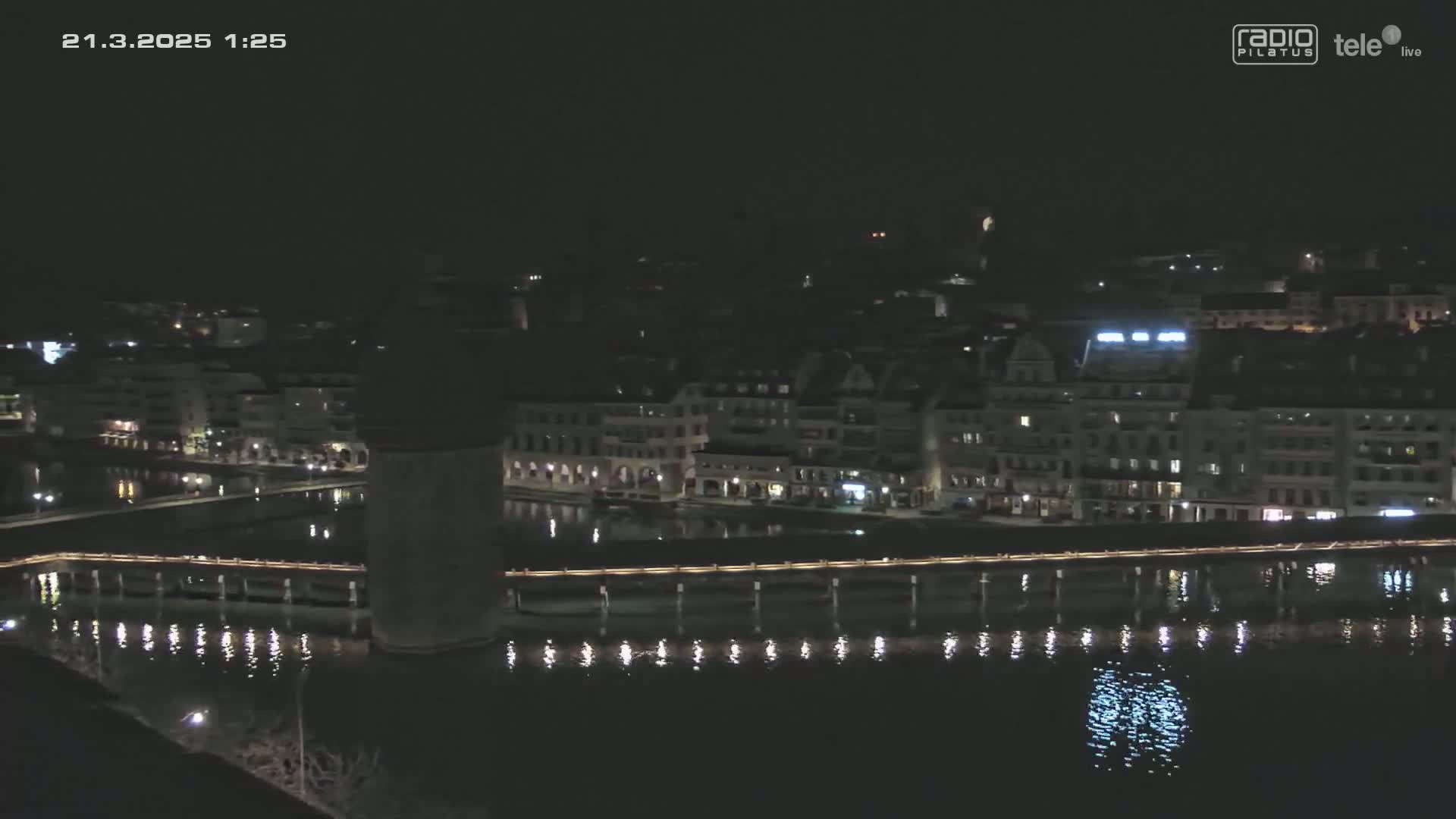 A nighttime view of a city's waterfront, showing buildings illuminated along a calm body of water with a bridge and tower.