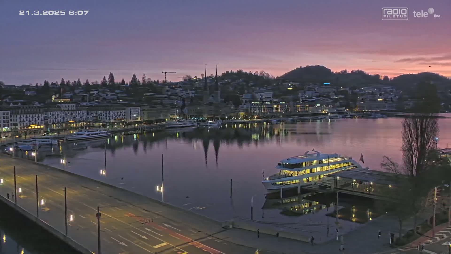 A city waterfront at twilight, with a large boat docked near a bridge and calm water reflecting the city lights under a purplish-pink sky.