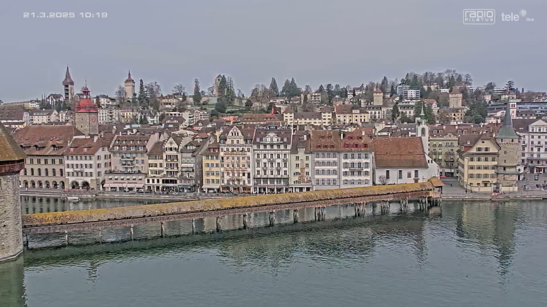 A long, covered wooden bridge spans a calm body of water in front of a European town under an overcast sky.