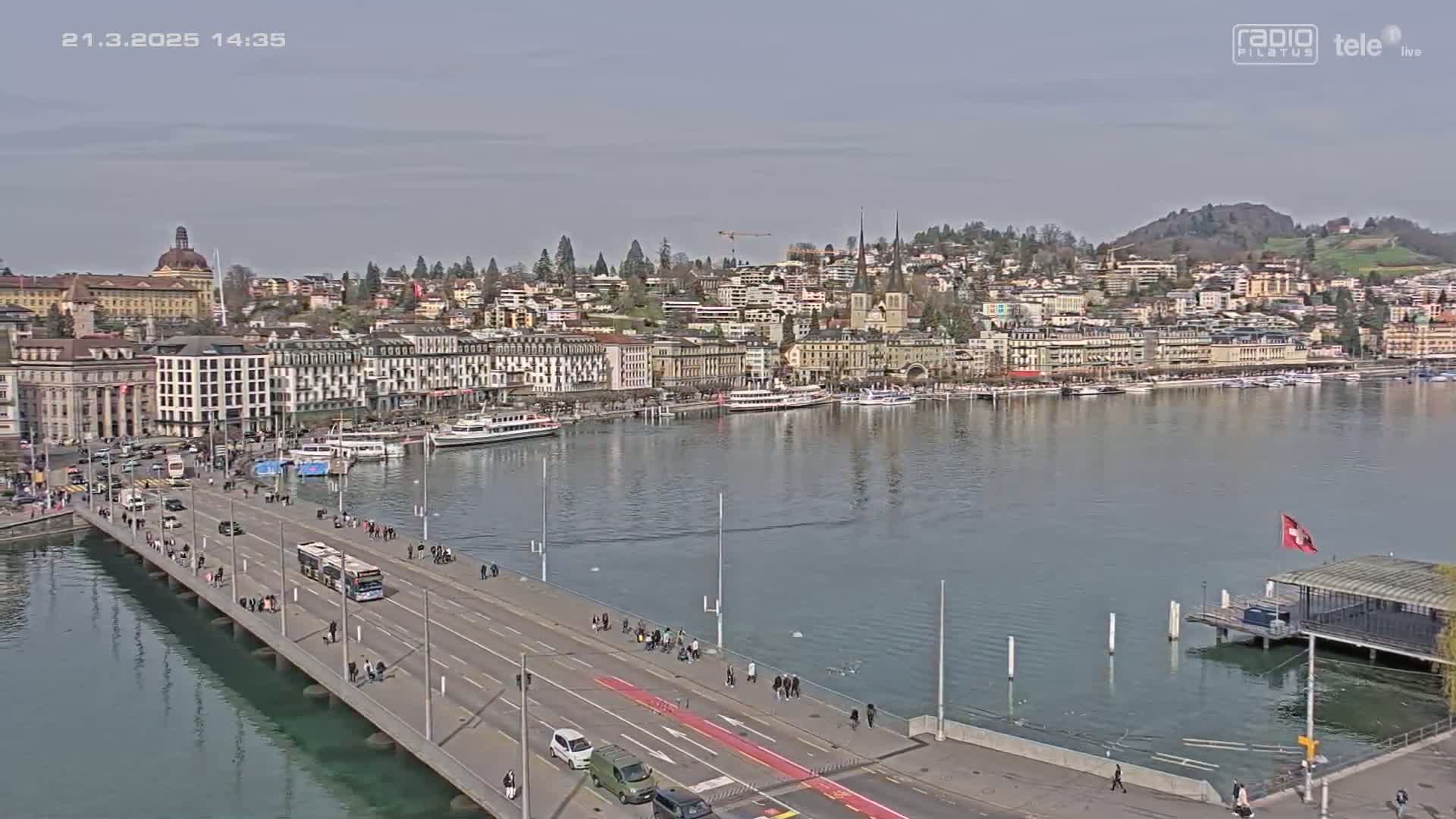 A bridge stretches across calm water, leading to a waterfront city with numerous buildings and docked boats under a mostly clear sky.