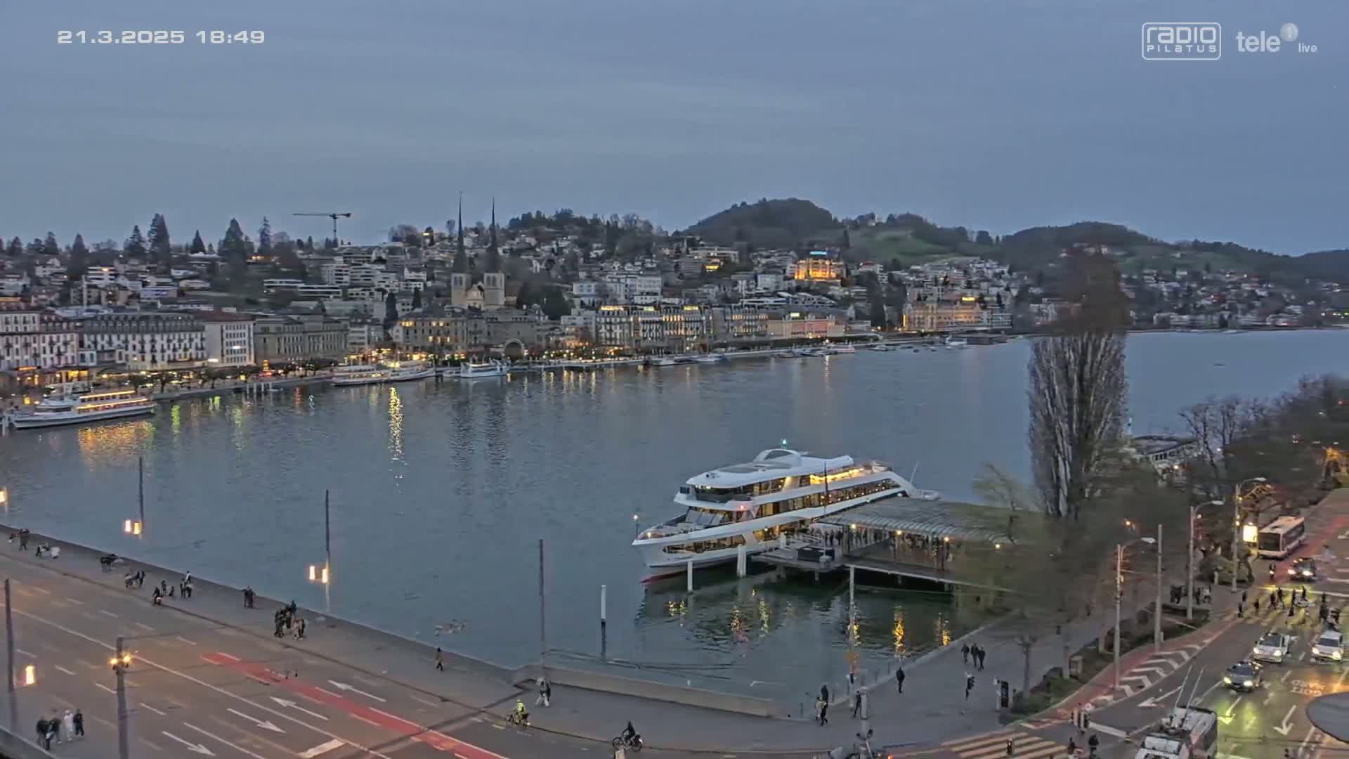 A large passenger ship is docked at a waterfront city at dusk under a twilight sky.