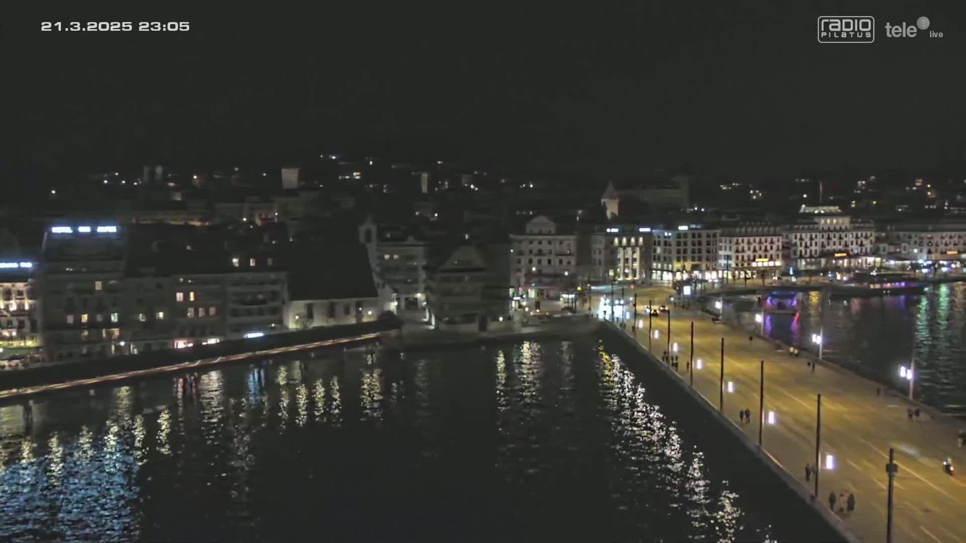 A nighttime, aerial view of a city's waterfront, showing a bridge lined with lights extending from a building-filled shoreline across calm, reflective water.