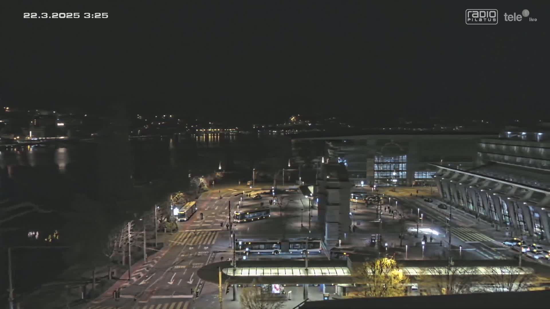 A nighttime aerial view shows a city transportation hub with trams and buses, near a body of water under a clear, dark sky.