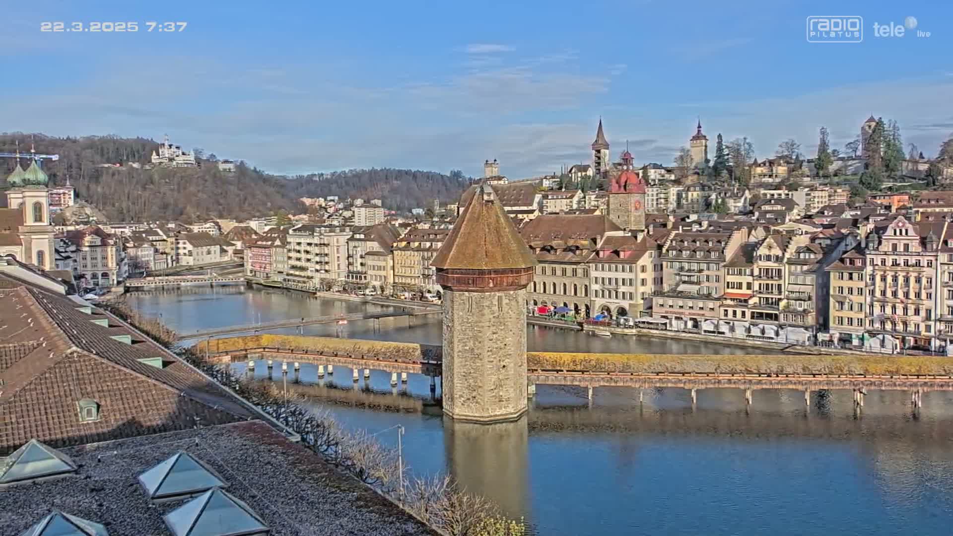 A stone tower stands in the middle of a river, with a covered bridge extending to either side, surrounded by a city of European-style buildings under a clear, sunny sky.