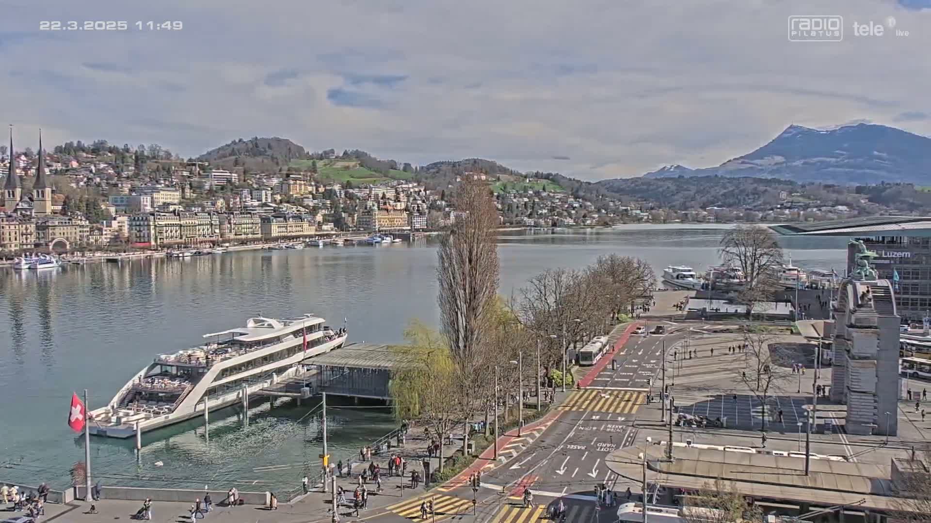 A lakeside city on a partly cloudy day, with a large passenger ship docked near a waterfront plaza and several smaller boats on the water.