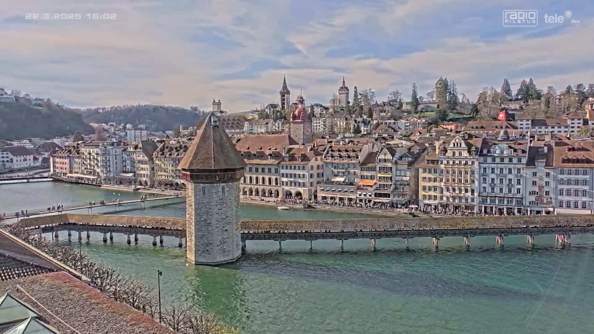 A stone water tower stands at the end of a long bridge extending over a body of green water, with a town of closely-packed buildings and church steeples visible in the background under a partly cloudy sky.