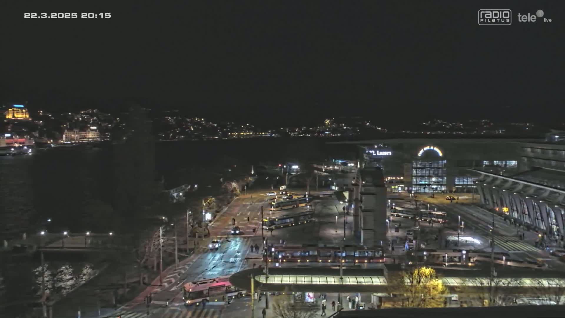 A nighttime aerial view of a city transportation hub with trams and buses, situated near a body of water and a city skyline under a clear, dark sky.
