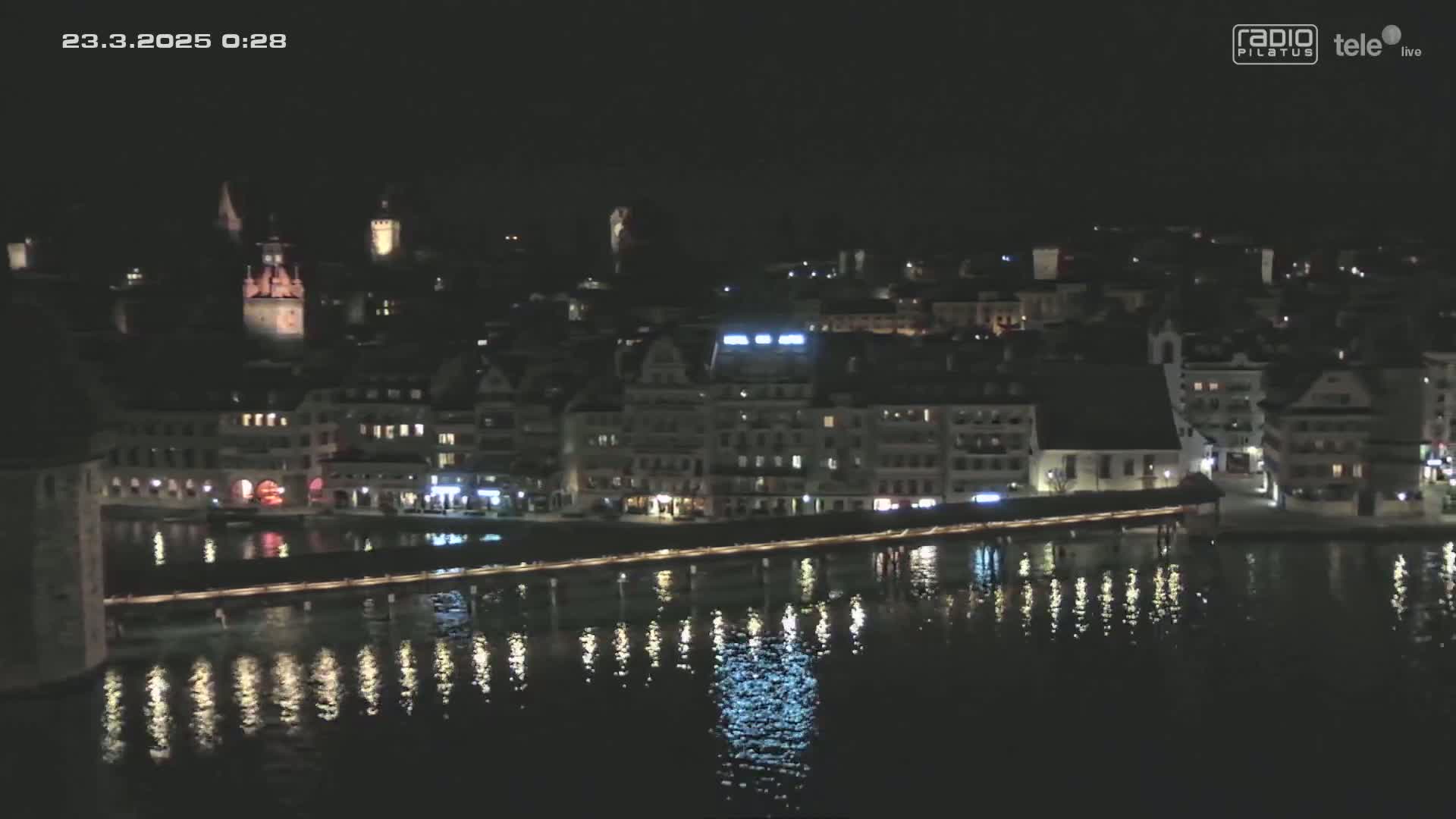 A nighttime view of a city with buildings lit along a waterfront, reflecting in calm water.
