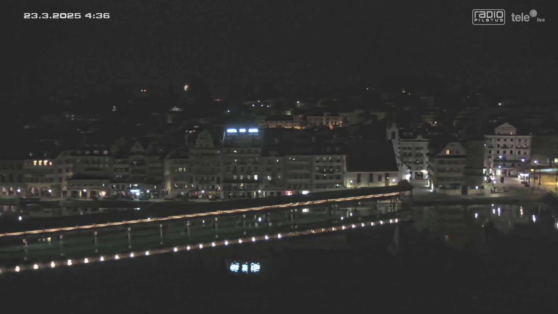 A nighttime, aerial view of a town situated on a body of water, showing buildings with lights and a bridge illuminated along the waterfront.