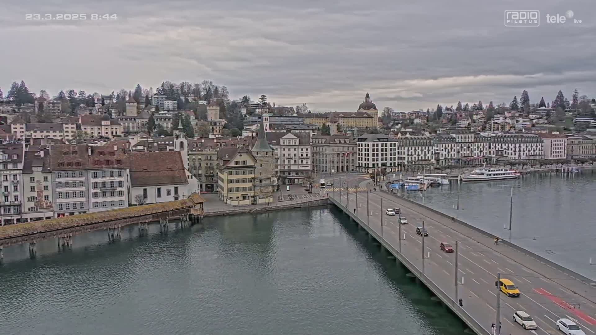A city with many buildings sits on a body of water under an overcast sky, a bridge crosses the water with cars driving on it.