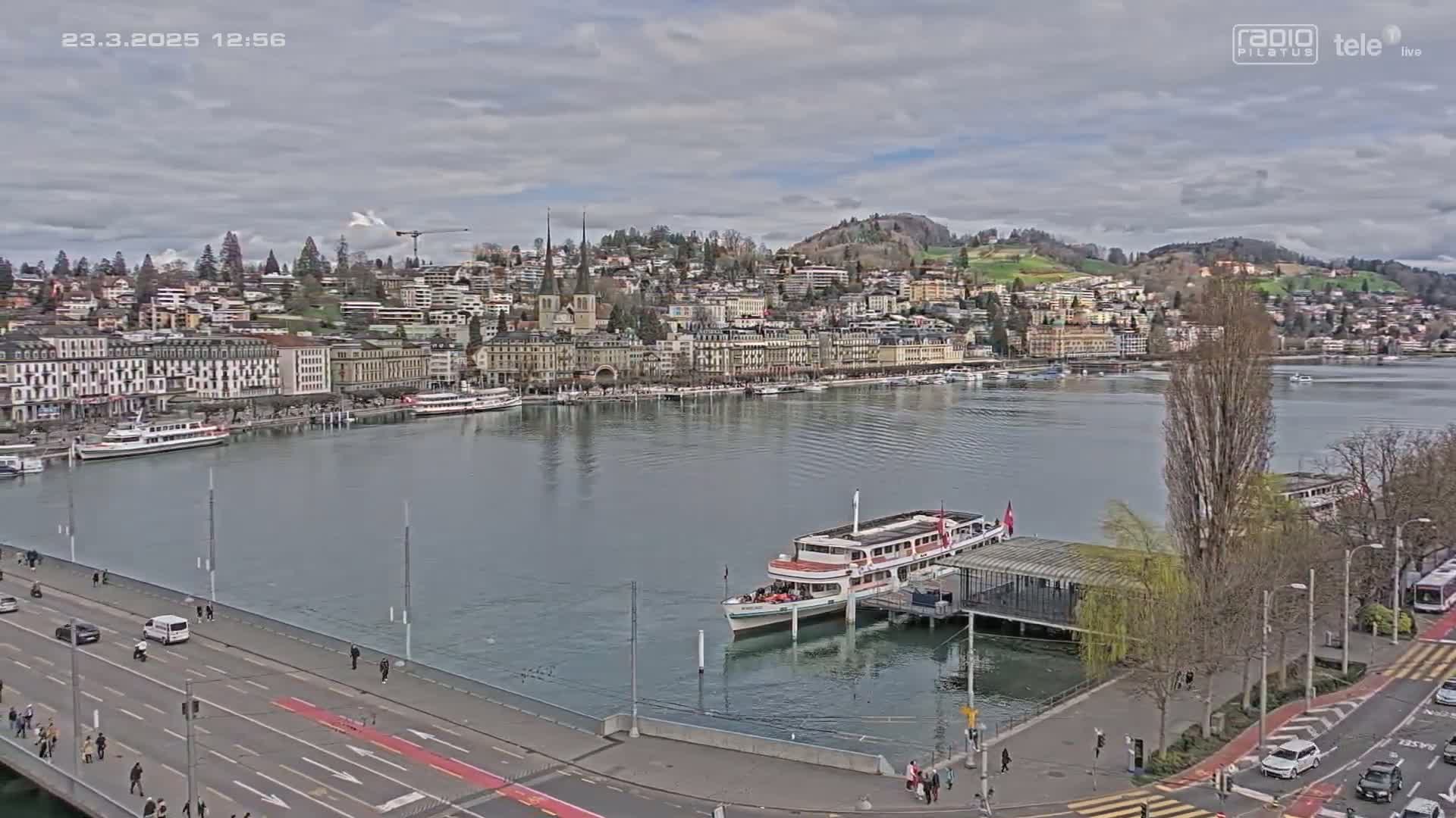 A city waterfront on a cloudy day shows a body of water with a docked passenger ship, a bridge with traffic, and buildings lining the shore.