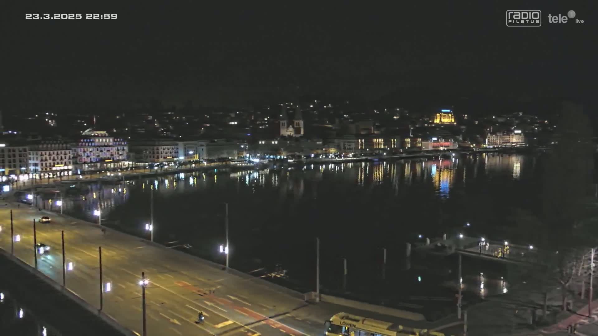 A nighttime, high-angle view of a city’s waterfront, featuring a bridge with sparse traffic, buildings illuminated along the shore, and calm water reflecting the city lights.