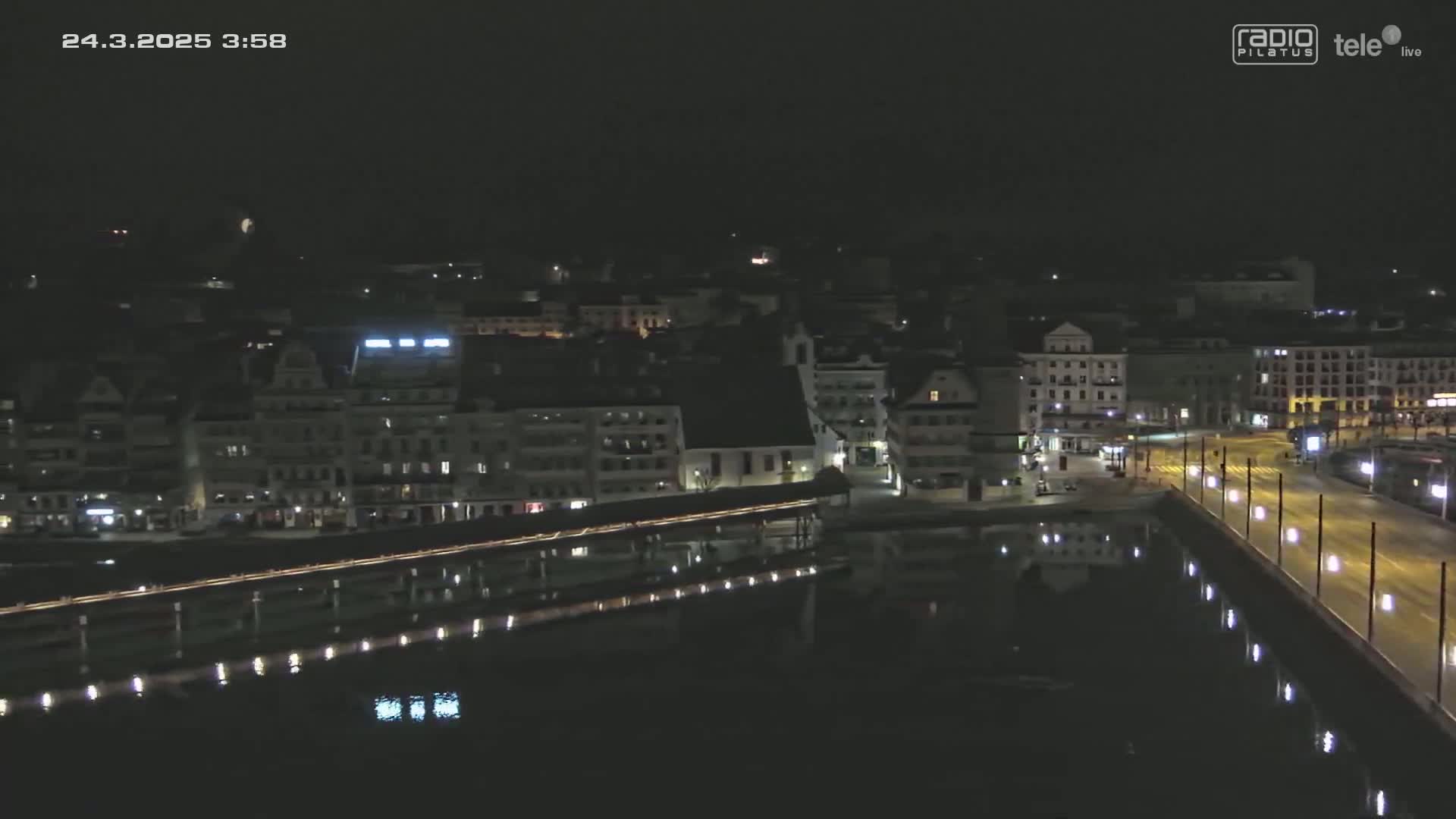 A nighttime, high-angle view shows a city with buildings and a bridge spanning a body of water, which reflects the lights of the city and bridge.