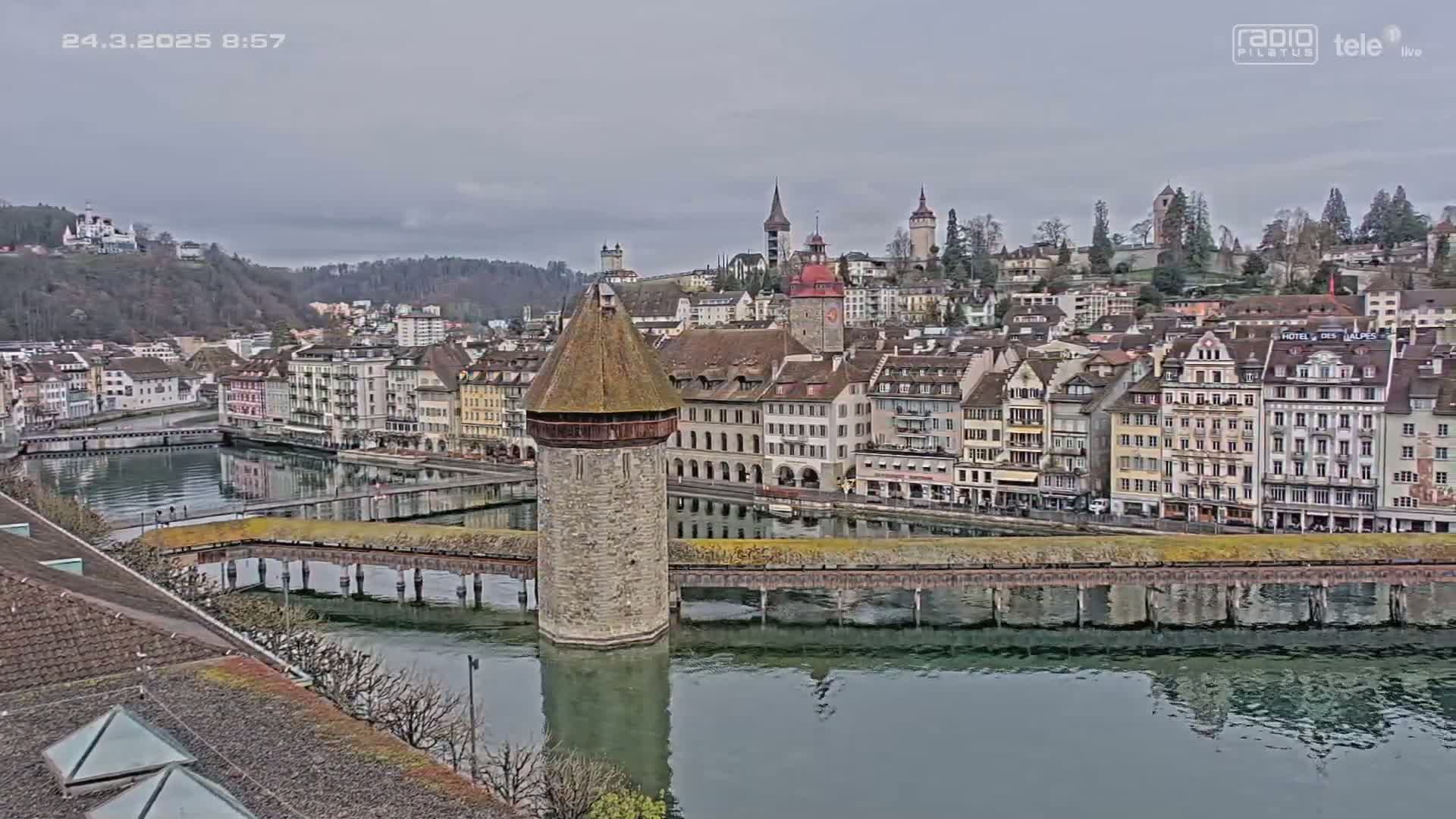 A stone water tower sits in a calm river, surrounded by a picturesque town of European-style buildings under a cloudy sky.