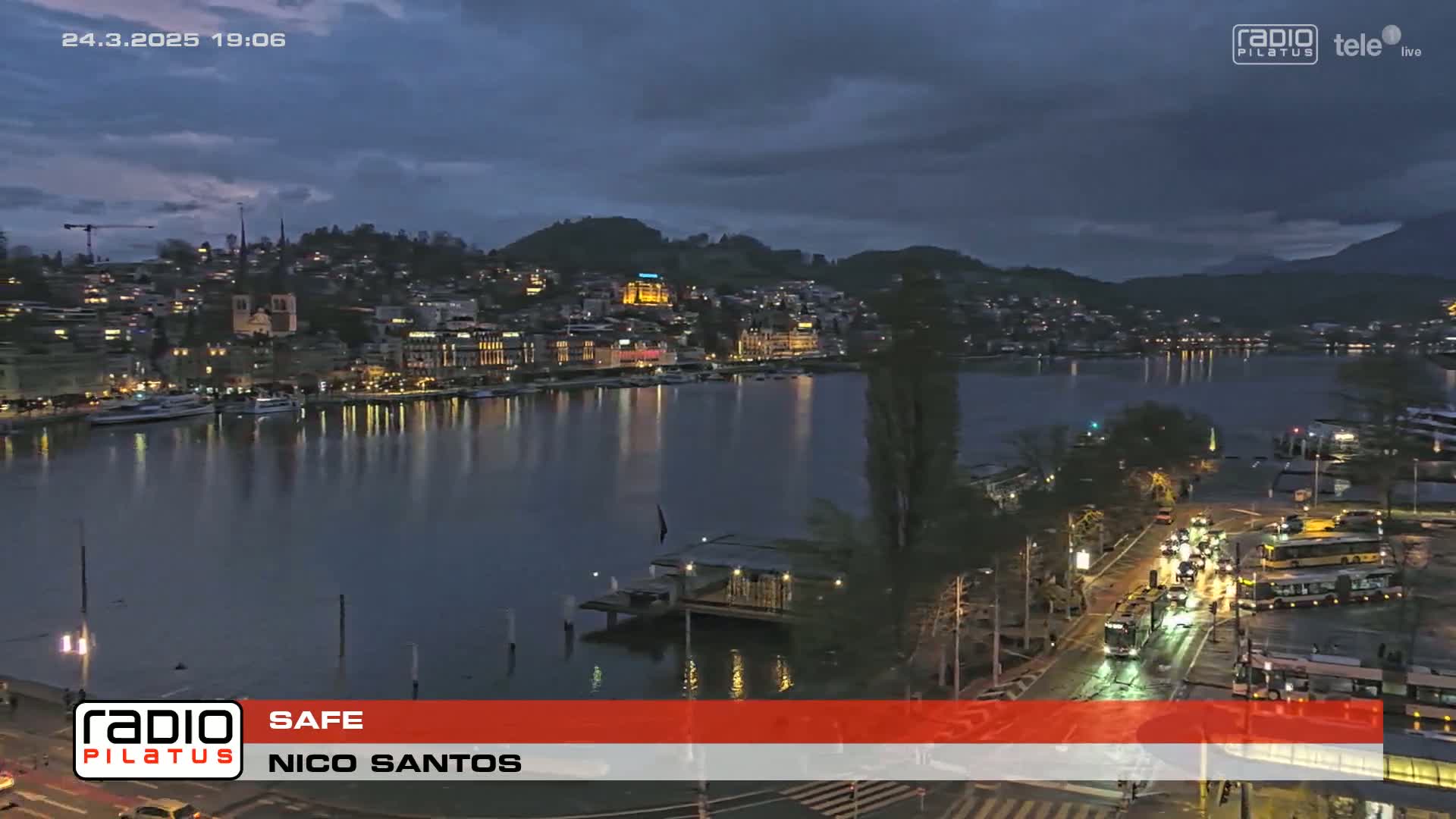 A city skyline on a waterfront at twilight under a partly cloudy sky, with boats moored on the water and traffic moving along a road beside it.