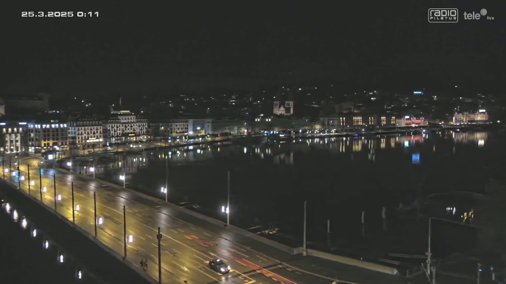 A nighttime, wet city scene shows a bridge with vehicles driving on it, and a calm body of water reflecting the city lights across from a waterfront lined with buildings.