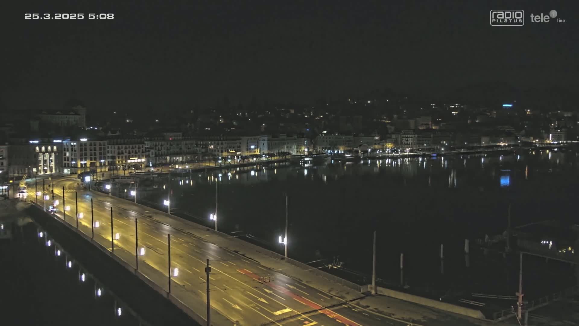A nighttime, high-angle view shows a bridge with traffic crossing over a calm body of water, with a city illuminated on the opposite shore.