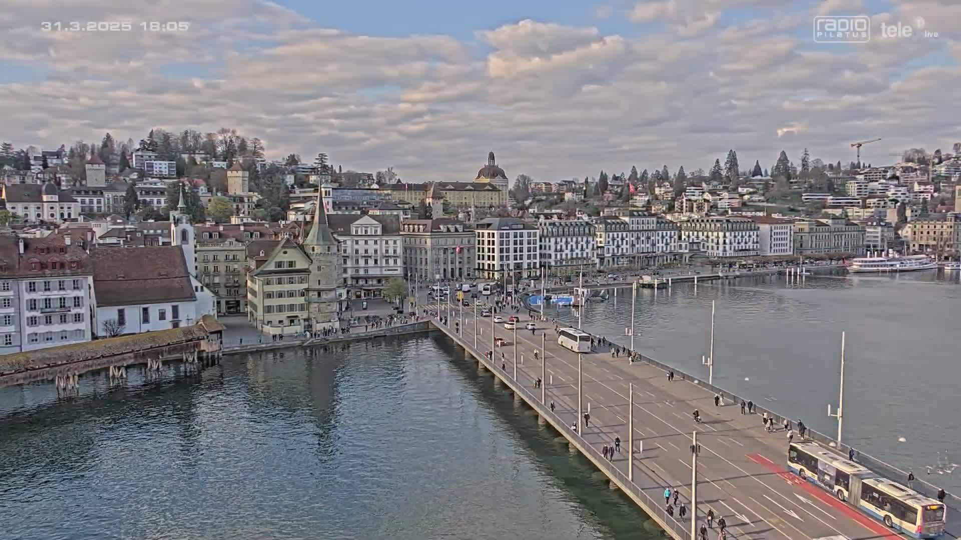 A bridge over a calm body of water leads to a city with many buildings under a partly cloudy sky.