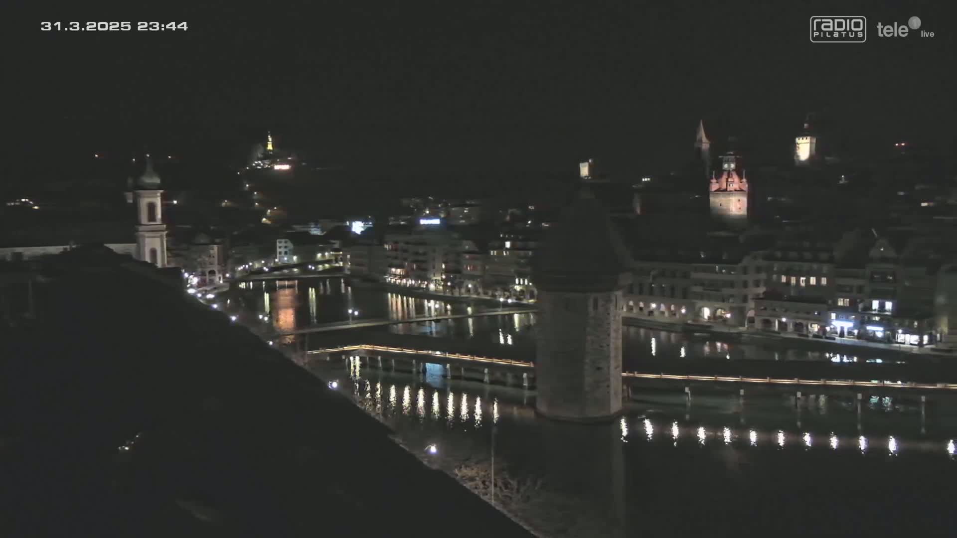 A nighttime view of a city beside a body of water, showing buildings with lights on and a bridge spanning the water.