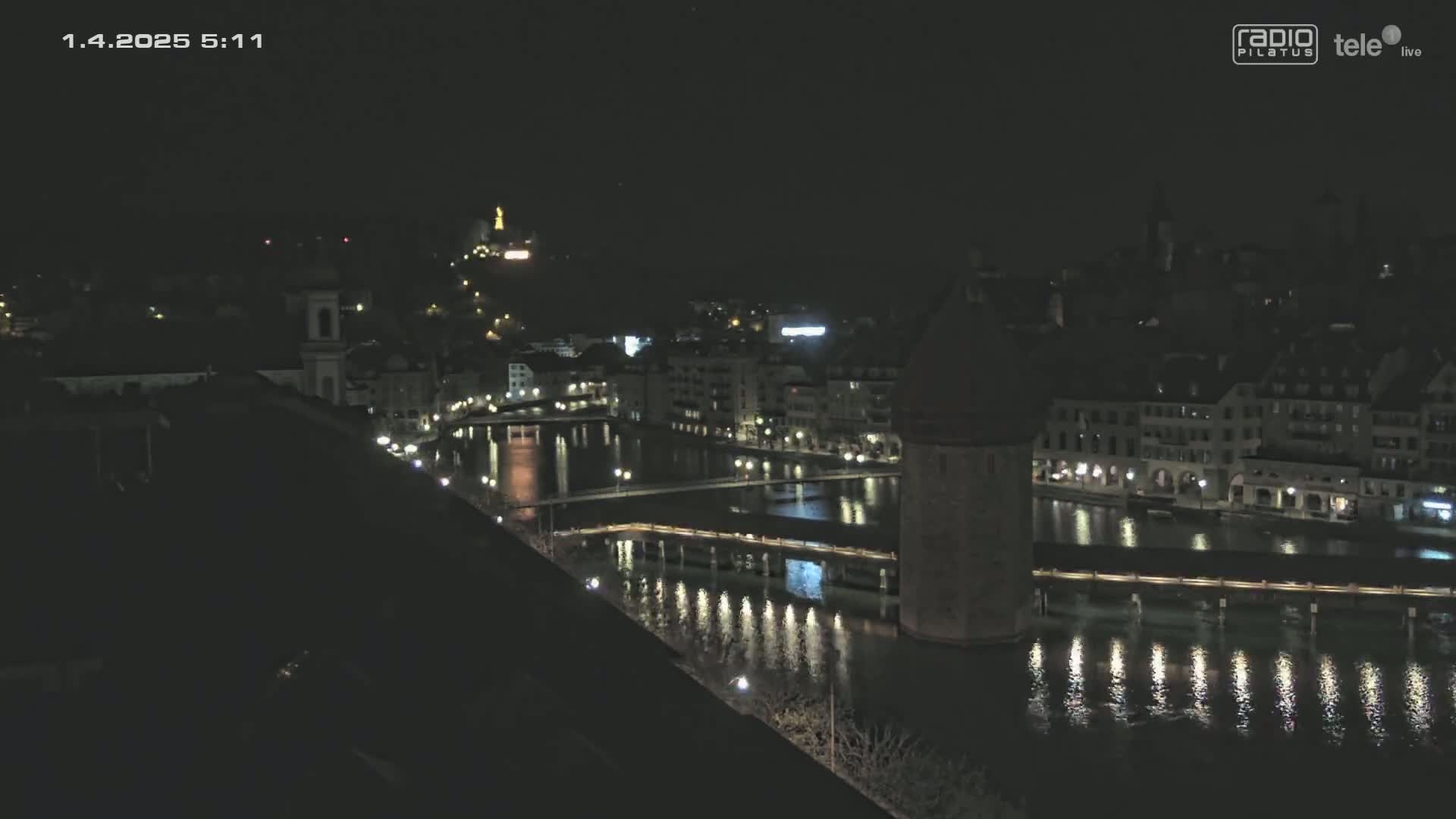 A nighttime view of a city with a river reflecting lights from buildings and bridges under a clear, dark sky.
