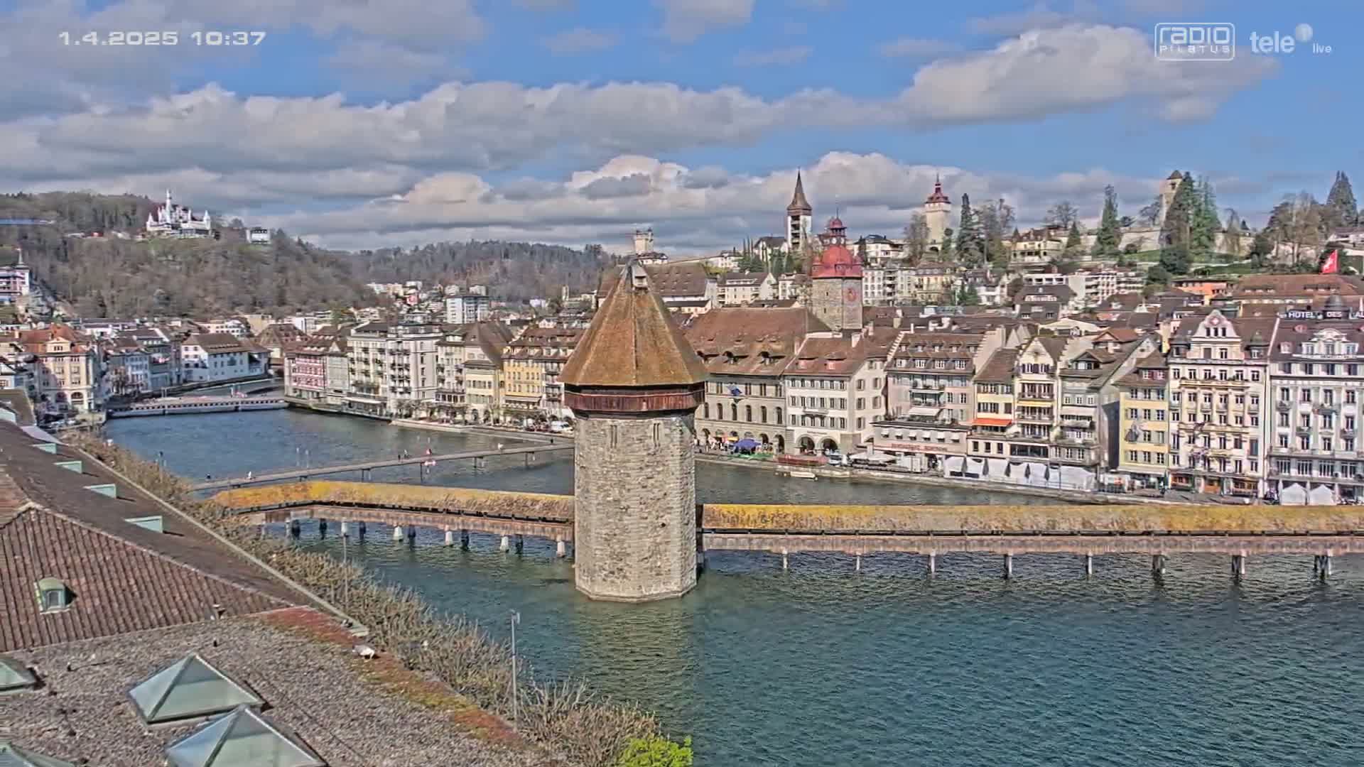 A stone tower stands in the middle of a covered wooden bridge spanning a river, with a collection of buildings lining the riverbanks under a partly cloudy sky.