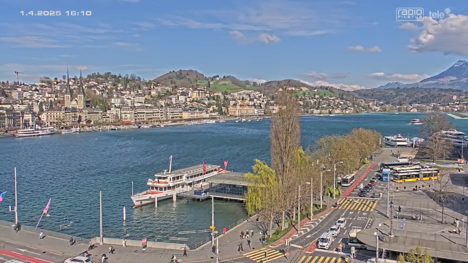 A lakefront city on a sunny day with a large passenger ship docked near a roadway with buses and cars.