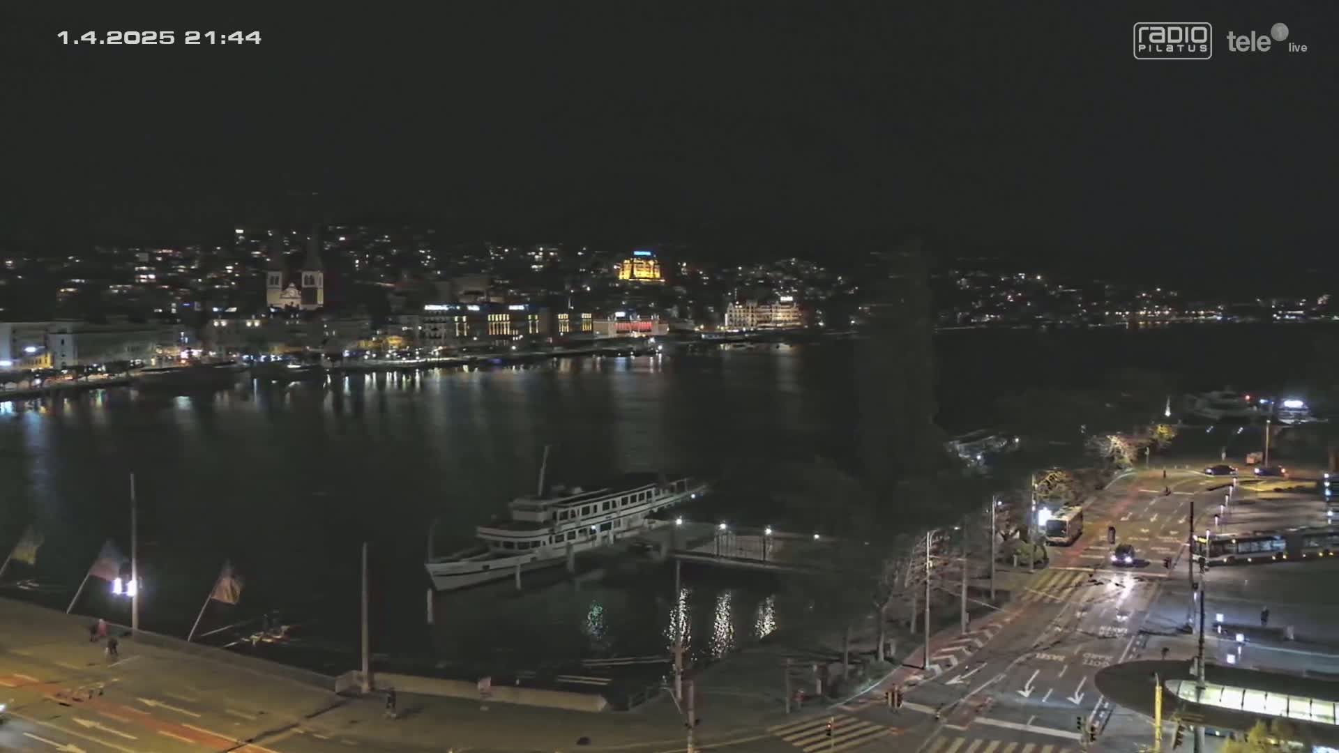 A nighttime view of a city waterfront with a docked boat and calm water, under clear skies.