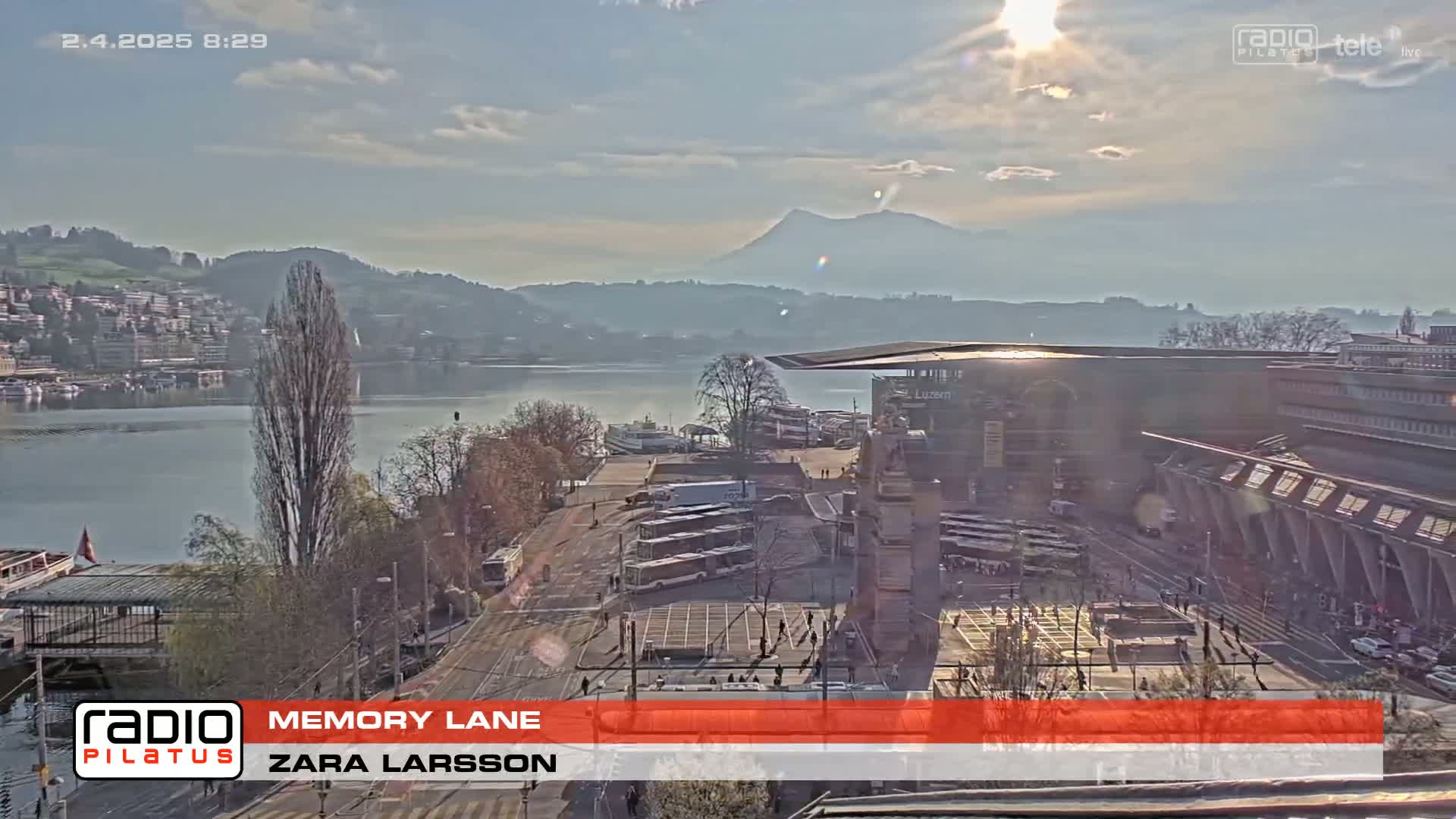 A sunny day overlooking a lake, city, and mountains, with a view of a transportation hub including trams and boats.