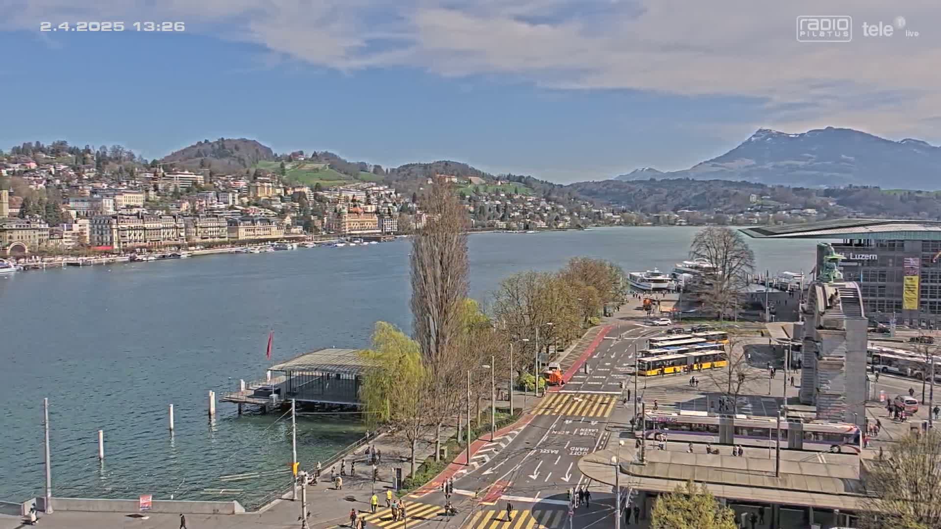 A sunny lakeside city scene shows a body of water with boats, buildings on a hill overlooking the water, a roadway with buses and trams, and mountains in the background.