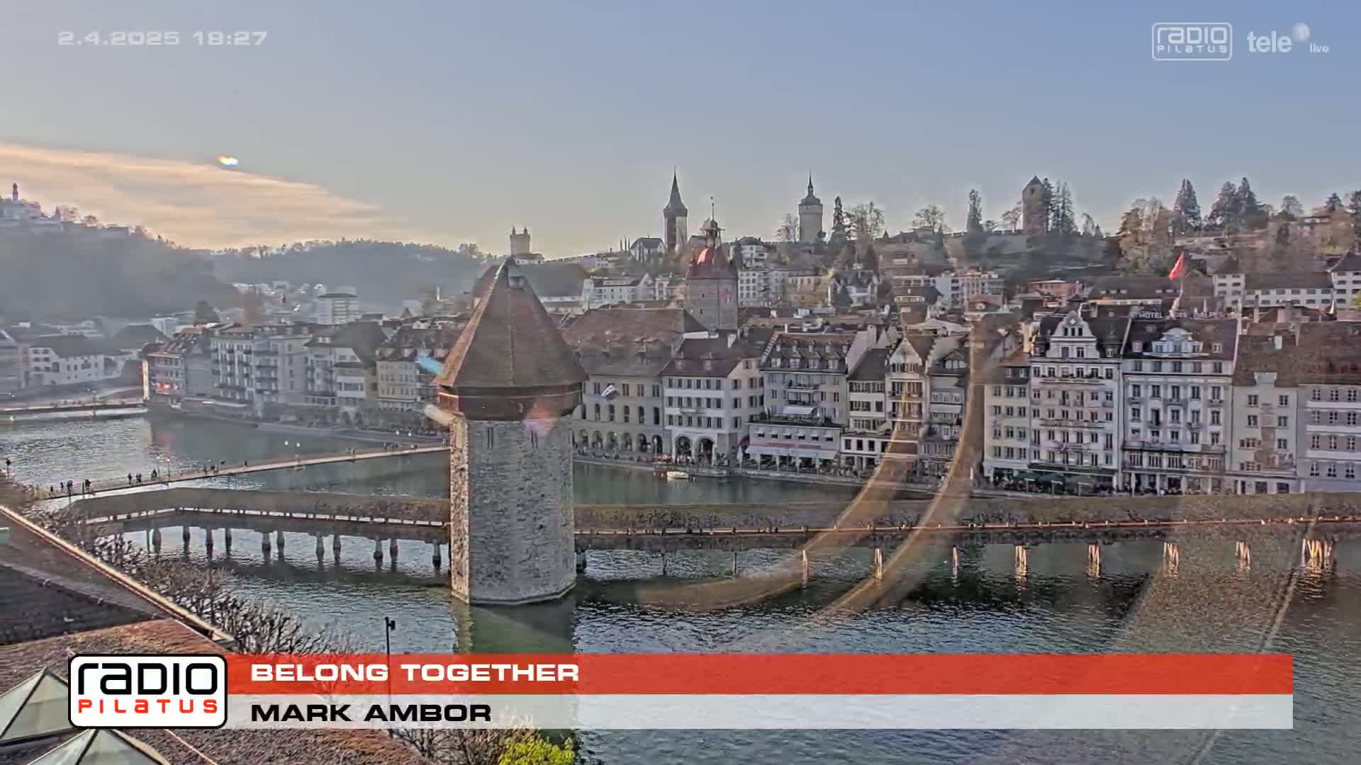 A stone water tower sits on a bridge over a calm body of water in front of a European town at sunset under a clear sky.