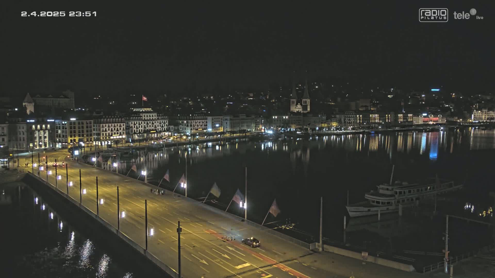 A nighttime view of a city's waterfront, showing a bridge with cars driving on it, a body of water reflecting city lights, and a docked boat.