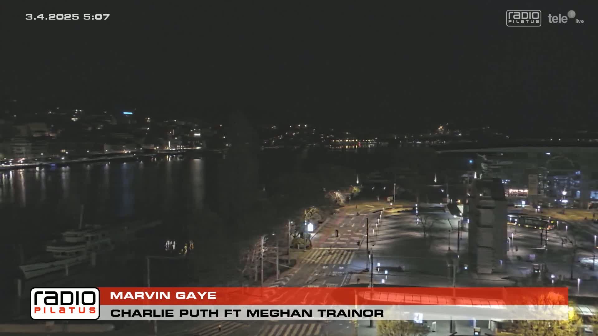 A nighttime, aerial view of a city waterfront with calm water, sparsely lit streets, and some docked boats.