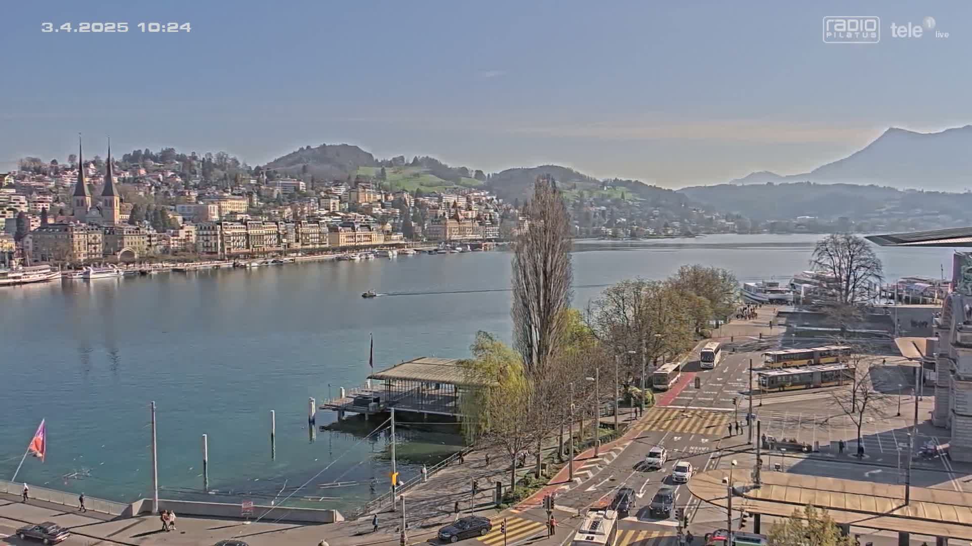 A waterfront city scene on a sunny day shows a calm lake with boats, a town of buildings on a hillside, and a paved area with cars and buses in the foreground.