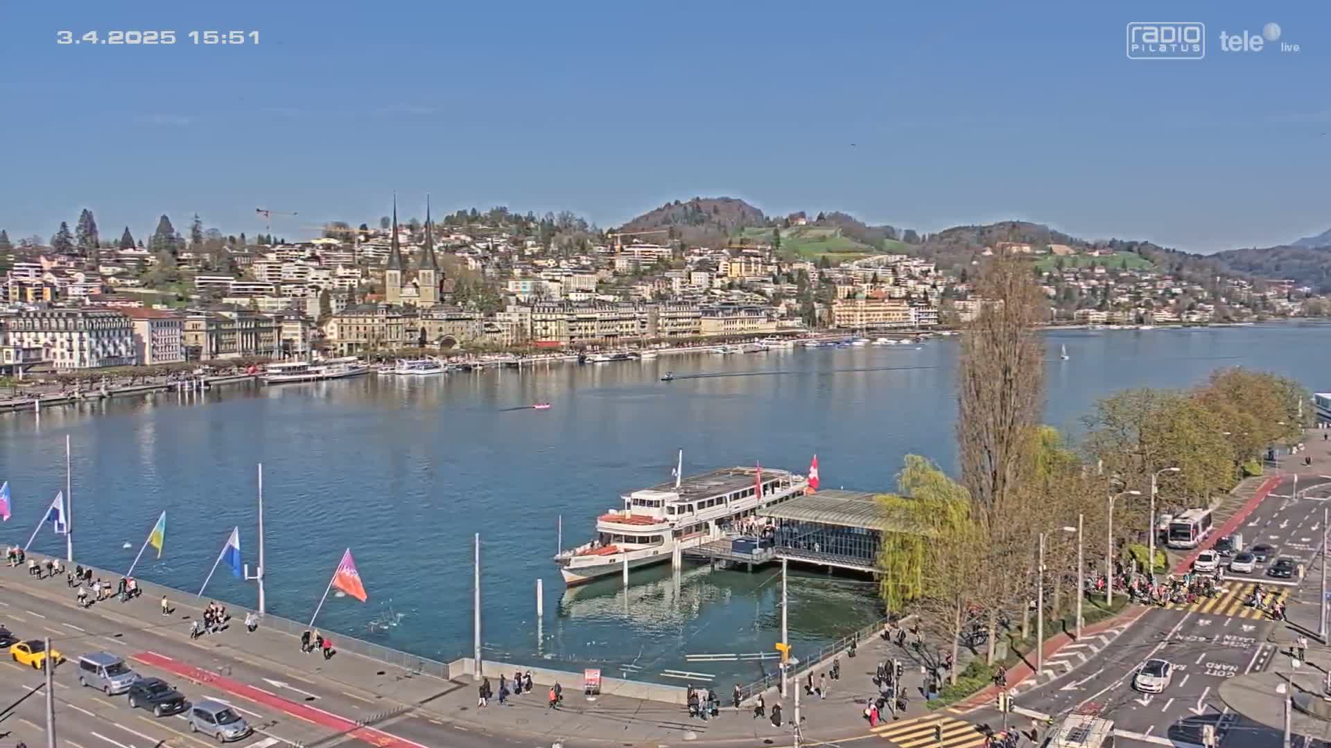 A lakefront town with buildings lining a calm lake under a clear blue sky, a passenger ship docked near a shoreline road with pedestrians and cars.