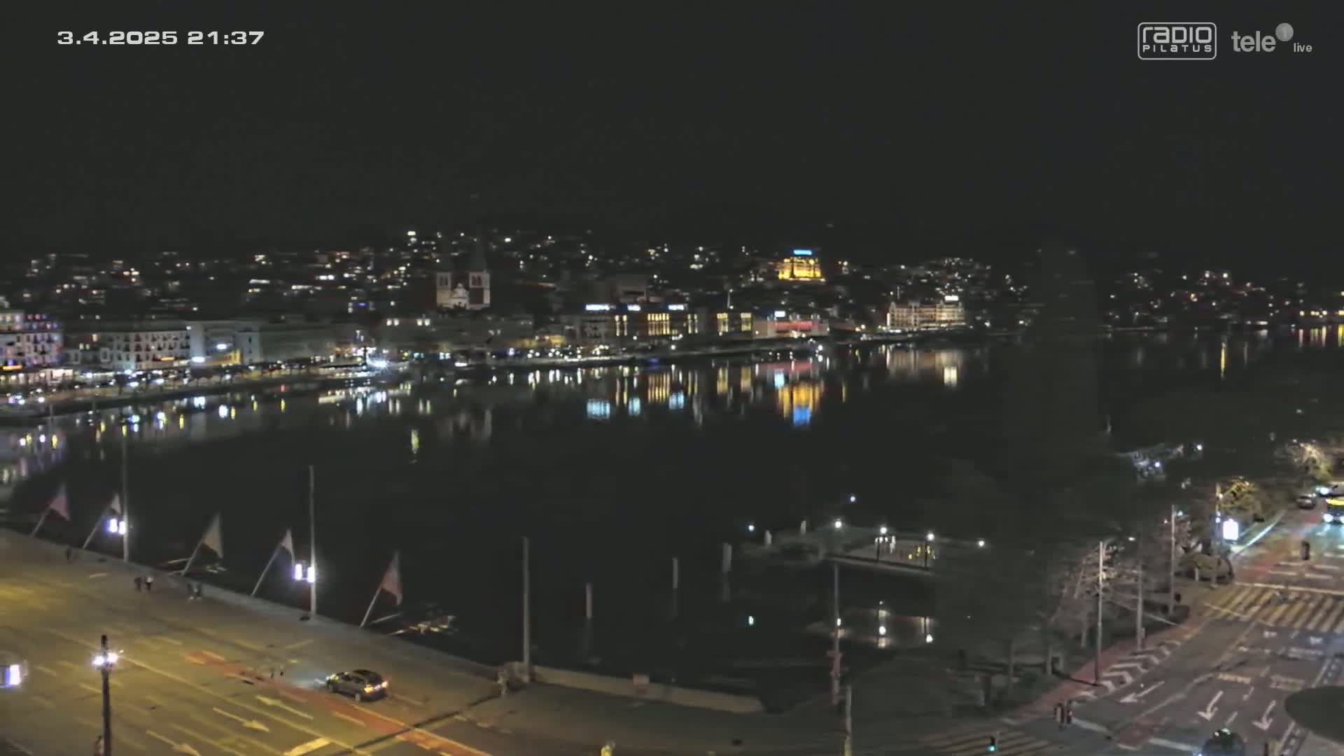 A nighttime view of a city waterfront, with lights reflecting on calm water under a clear, dark sky.