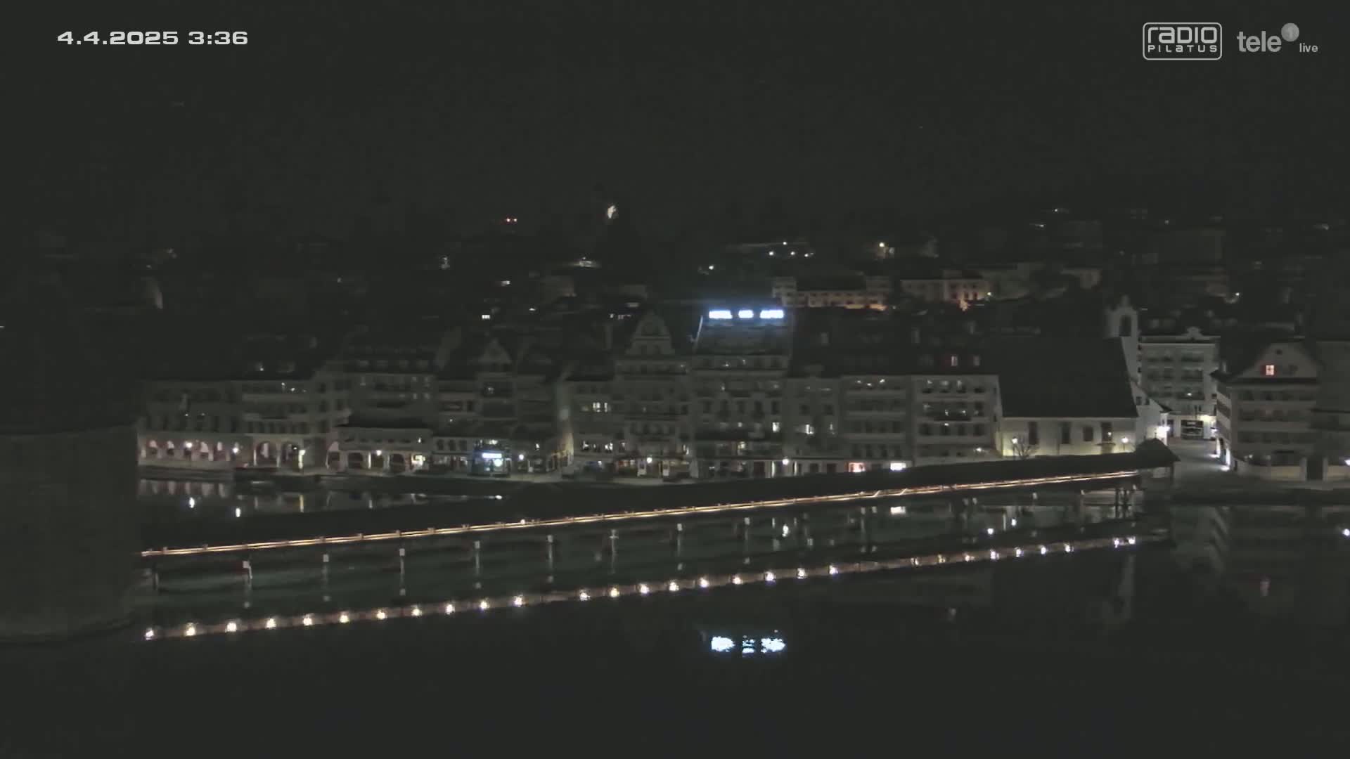A nighttime aerial view shows a town's waterfront with buildings lit along a body of water, a bridge, and a dark sky.