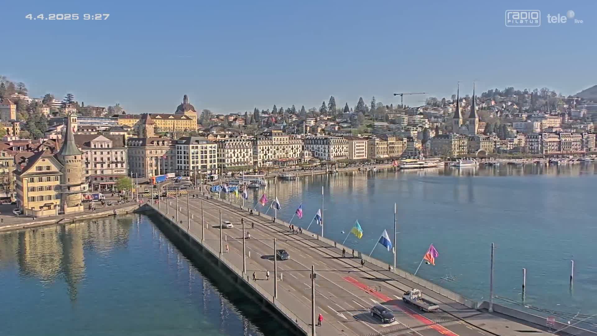 A long bridge spans a calm lake, leading to a city of numerous buildings under a clear blue sky.