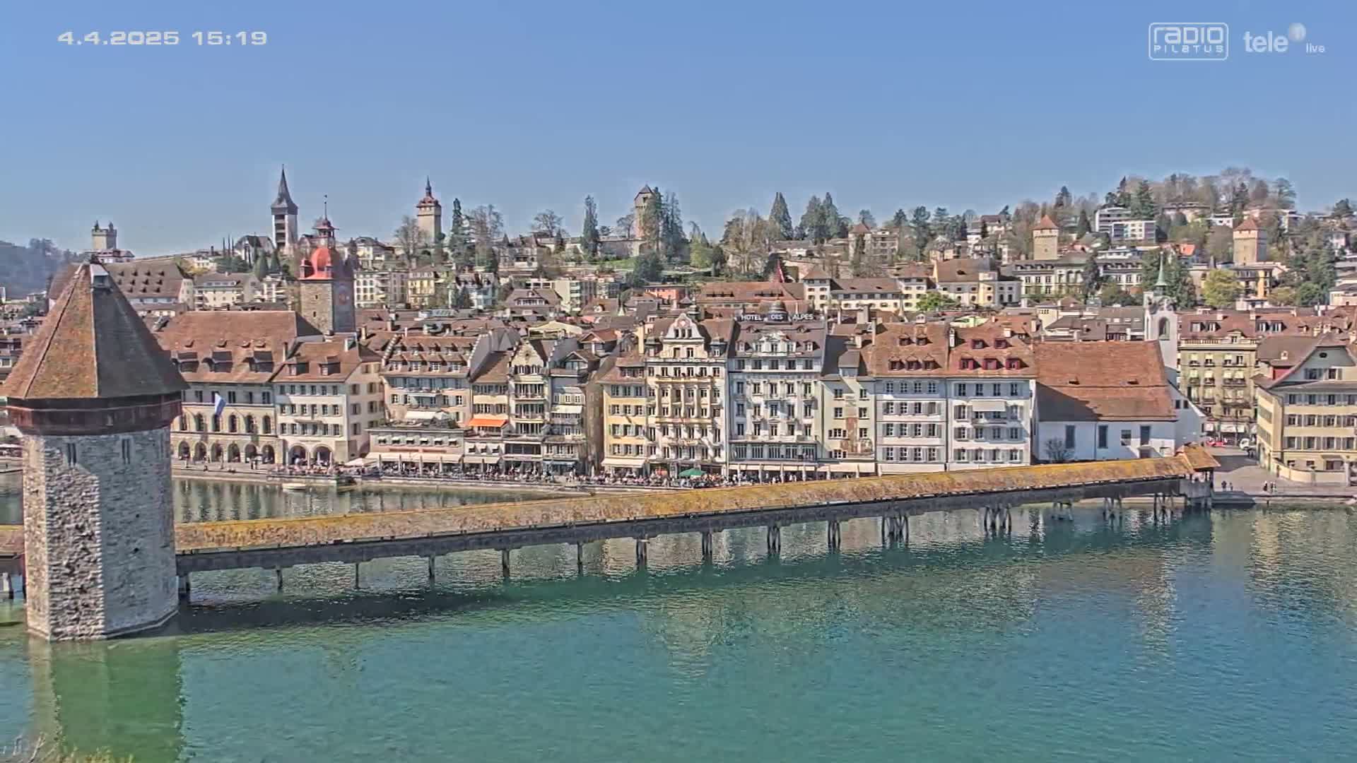 A wooden covered bridge spans a body of calm, blue-green water in front of a town of closely clustered buildings under a clear blue sky.