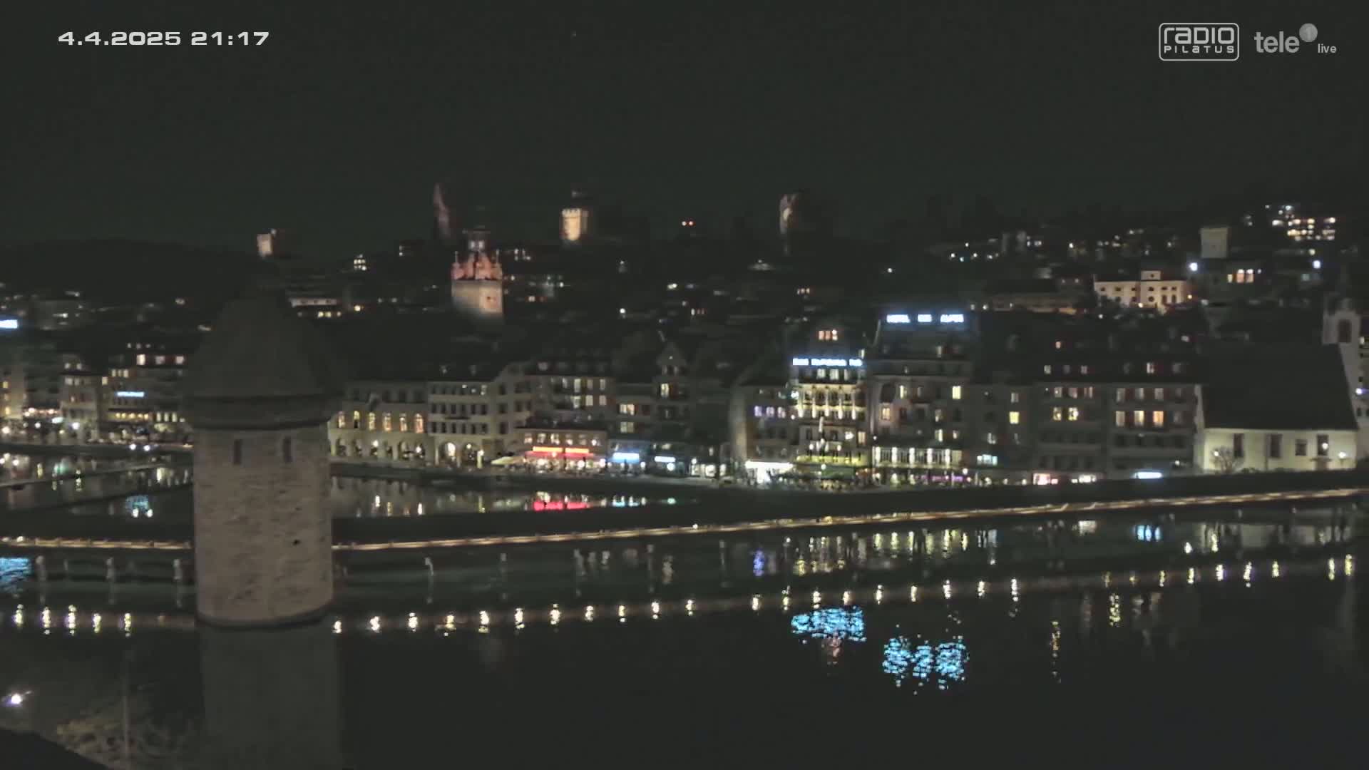 A nighttime view of a city with a river reflecting lights from buildings and a stone tower on a bridge.