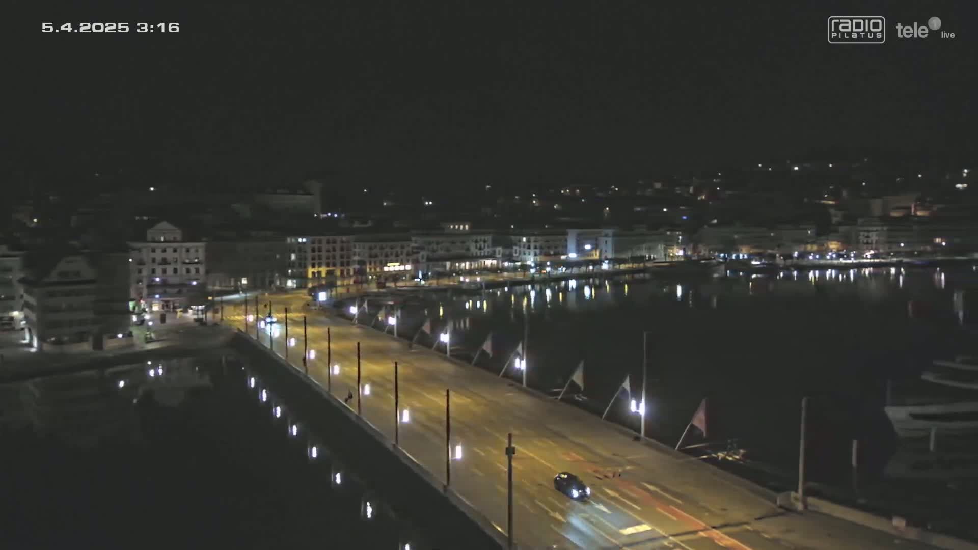 A nighttime view shows a bridge over a calm body of water, with a city's lights reflecting on the water and a few vehicles visible on the bridge.