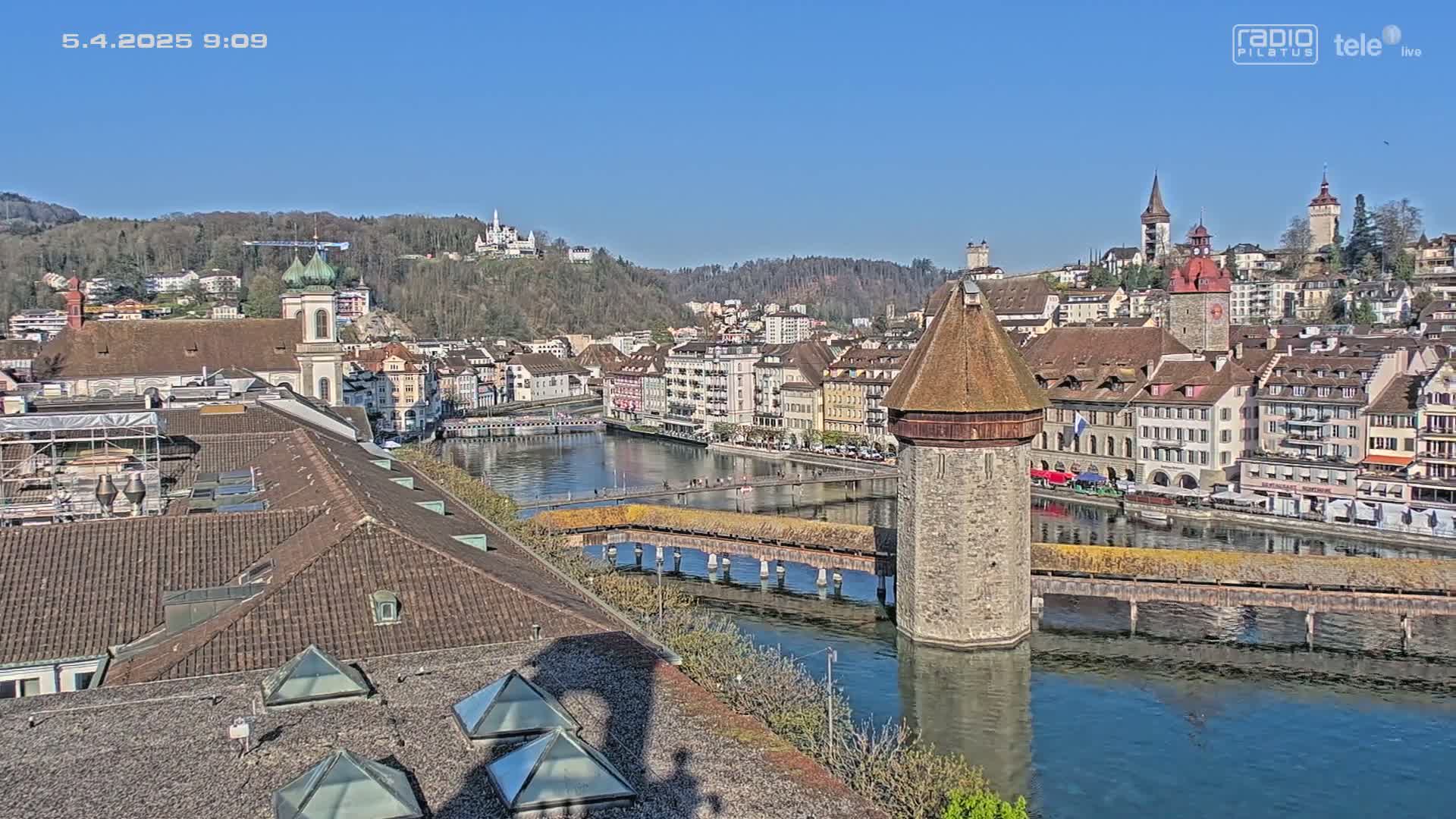 A sunny view of a European city with a river, a covered wooden bridge, and several historic buildings, including a stone tower and a church.