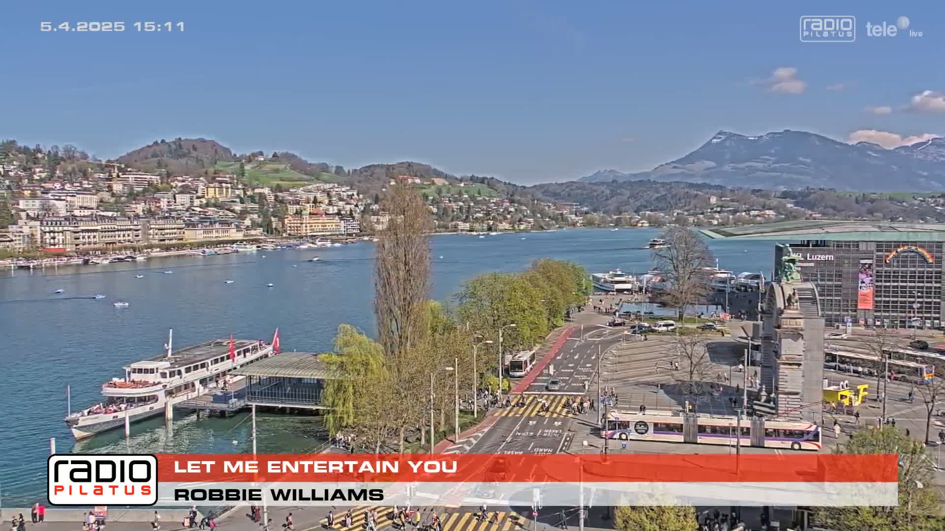 A sunny lakeside city scene shows a large body of water with boats and a town built along the shore, viewed from a high vantage point overlooking a roadway with pedestrians and trams.
