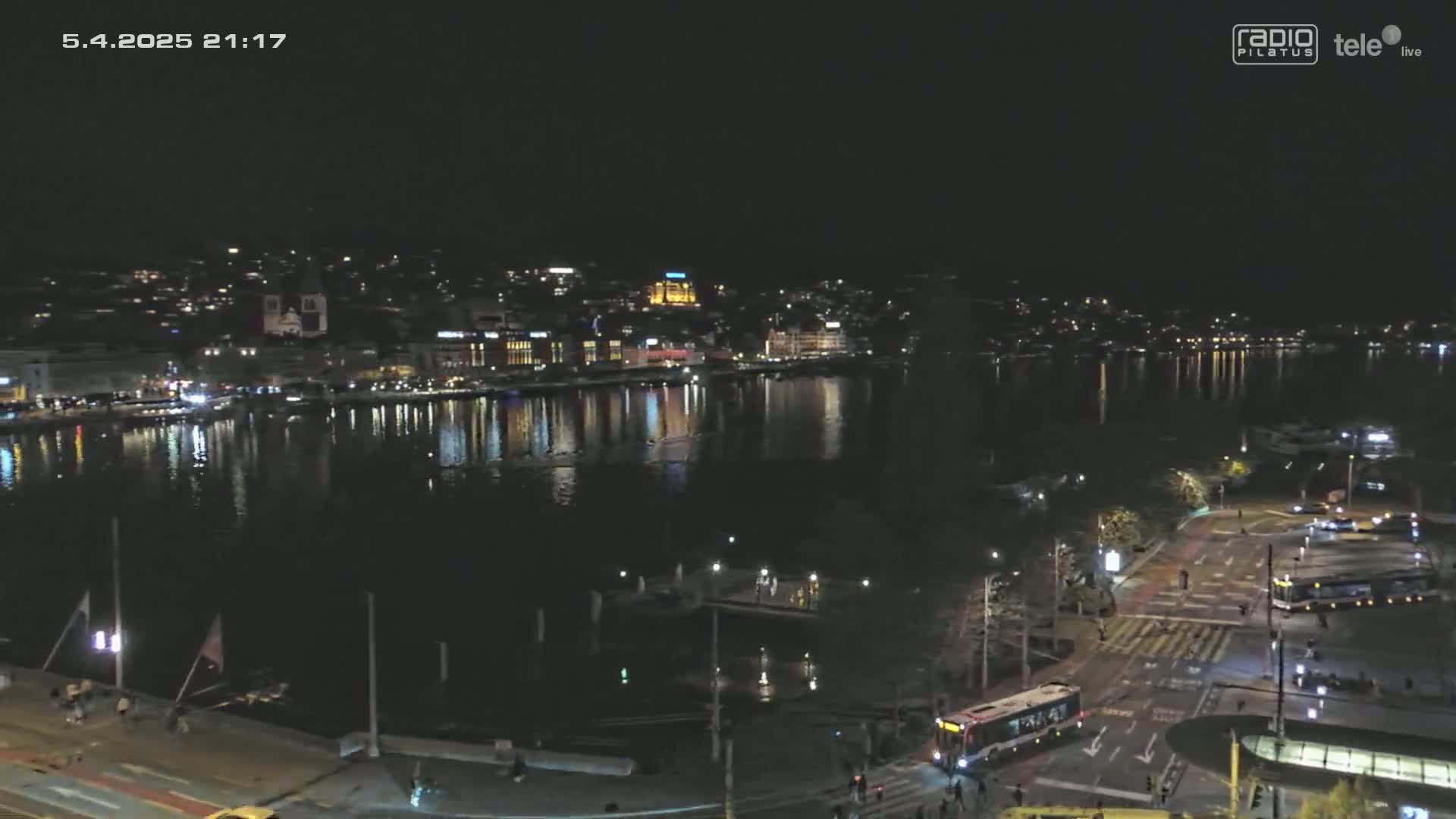 A nighttime view of a city waterfront with calm water reflecting city lights, under a clear sky.