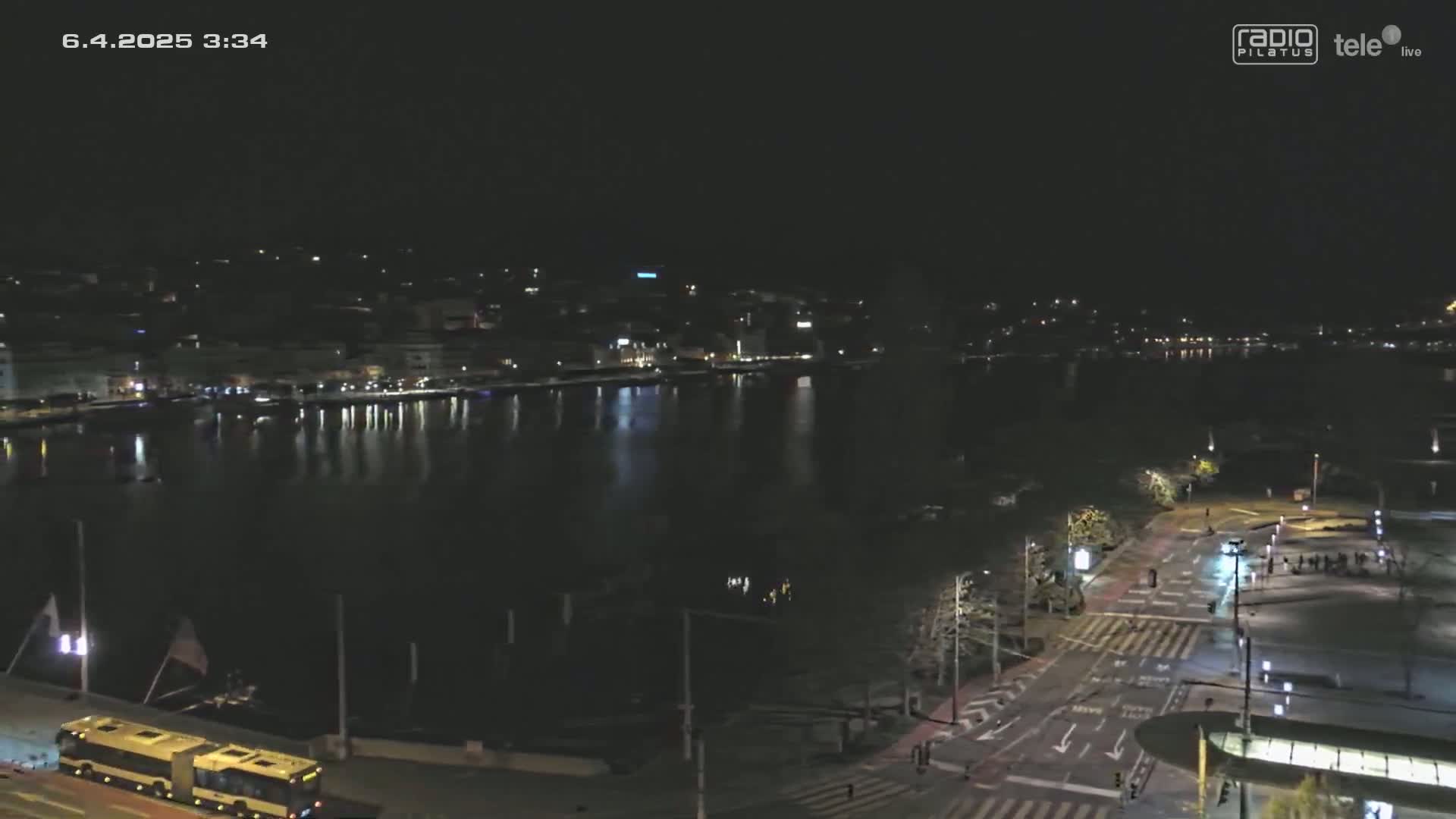 A nighttime, high-angle view shows a city waterfront at night, with a calm body of water reflecting city lights, and roads and a bus visible in the foreground.