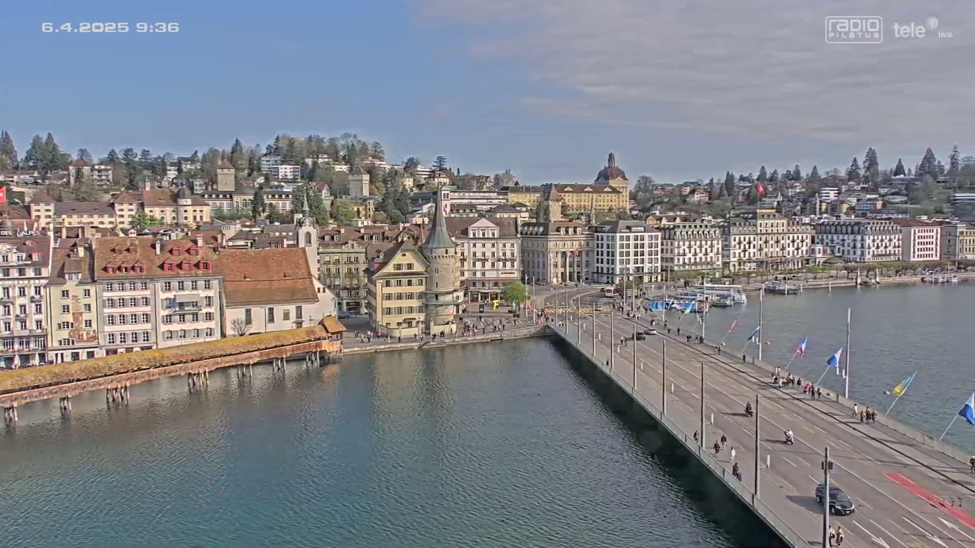 A sunny day shows a city by a lake with a long bridge extending from the waterfront to a collection of buildings, including a covered wooden bridge.