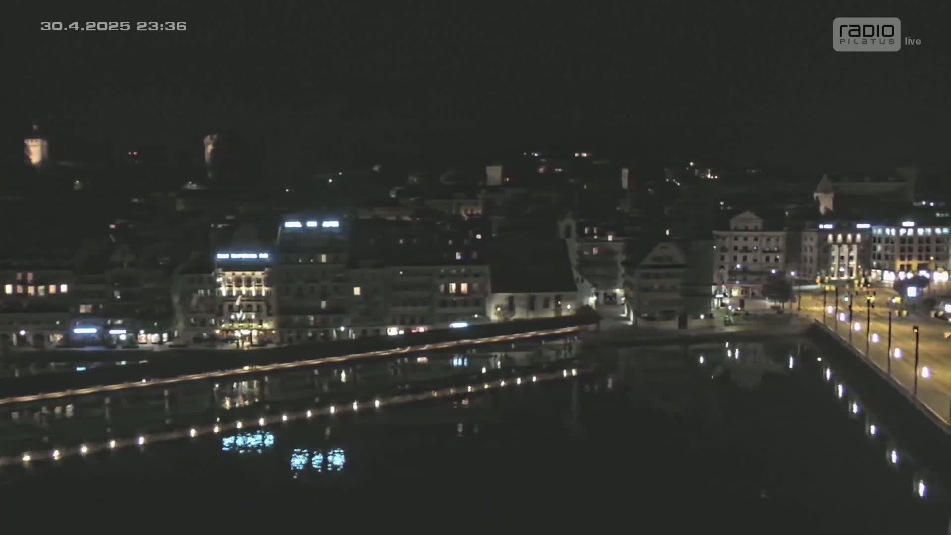 A nighttime, aerial view shows a city's waterfront with buildings illuminated along a calm body of water.