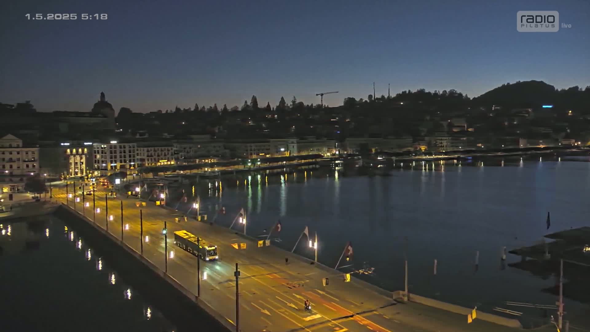 A nighttime, low-angle view shows a city waterfront with a bus driving along a bridge over calm water, city lights reflecting on the water's surface.