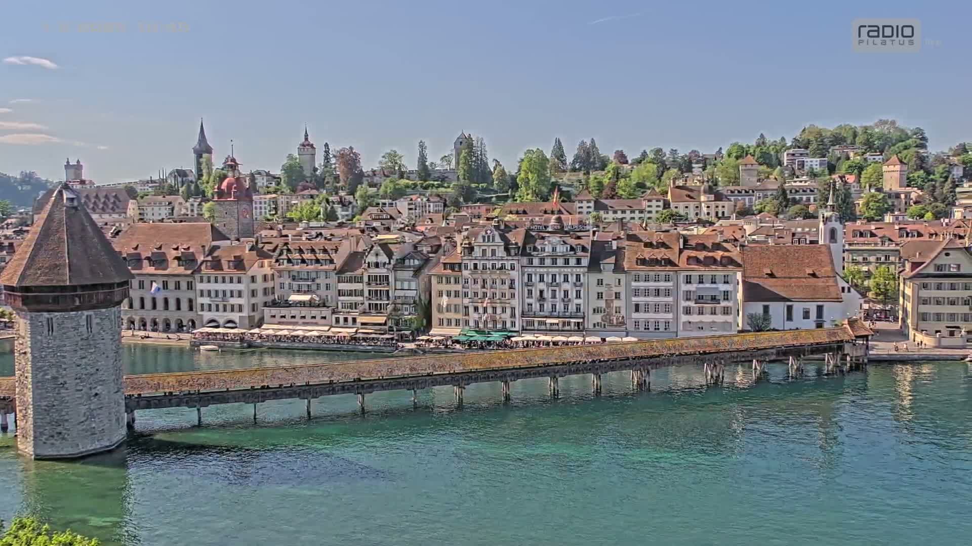 A wooden bridge stretches across a body of teal water, connecting to a town of European-style buildings on a sunny day.