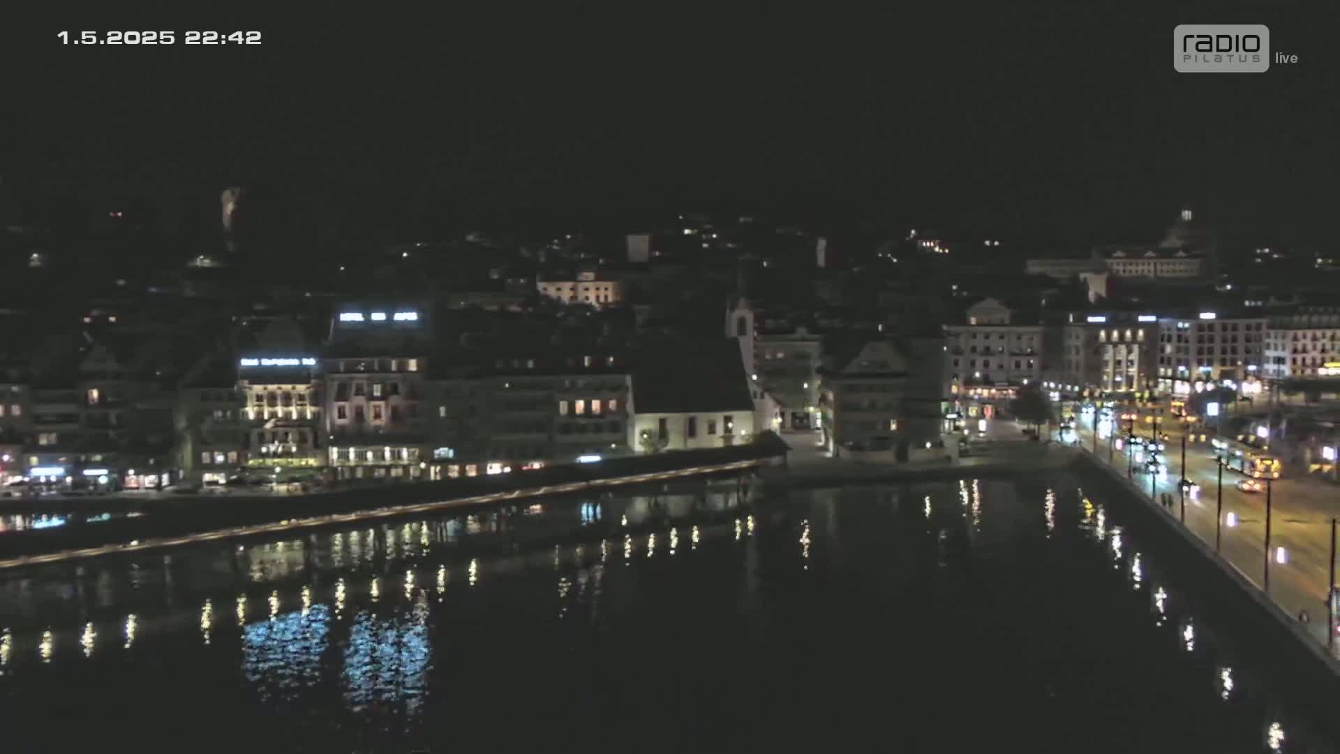 A nighttime, aerial view shows a city's waterfront with calm water reflecting the lights from buildings and streetlights along a road.