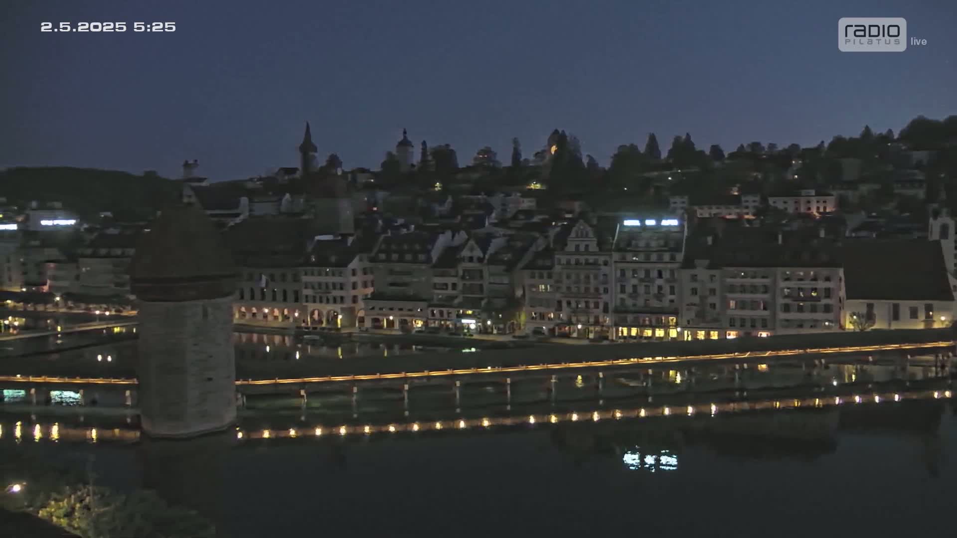 A nighttime view of a city with buildings illuminated along a waterfront, a stone tower visible near a bridge, under a dark, clear sky.